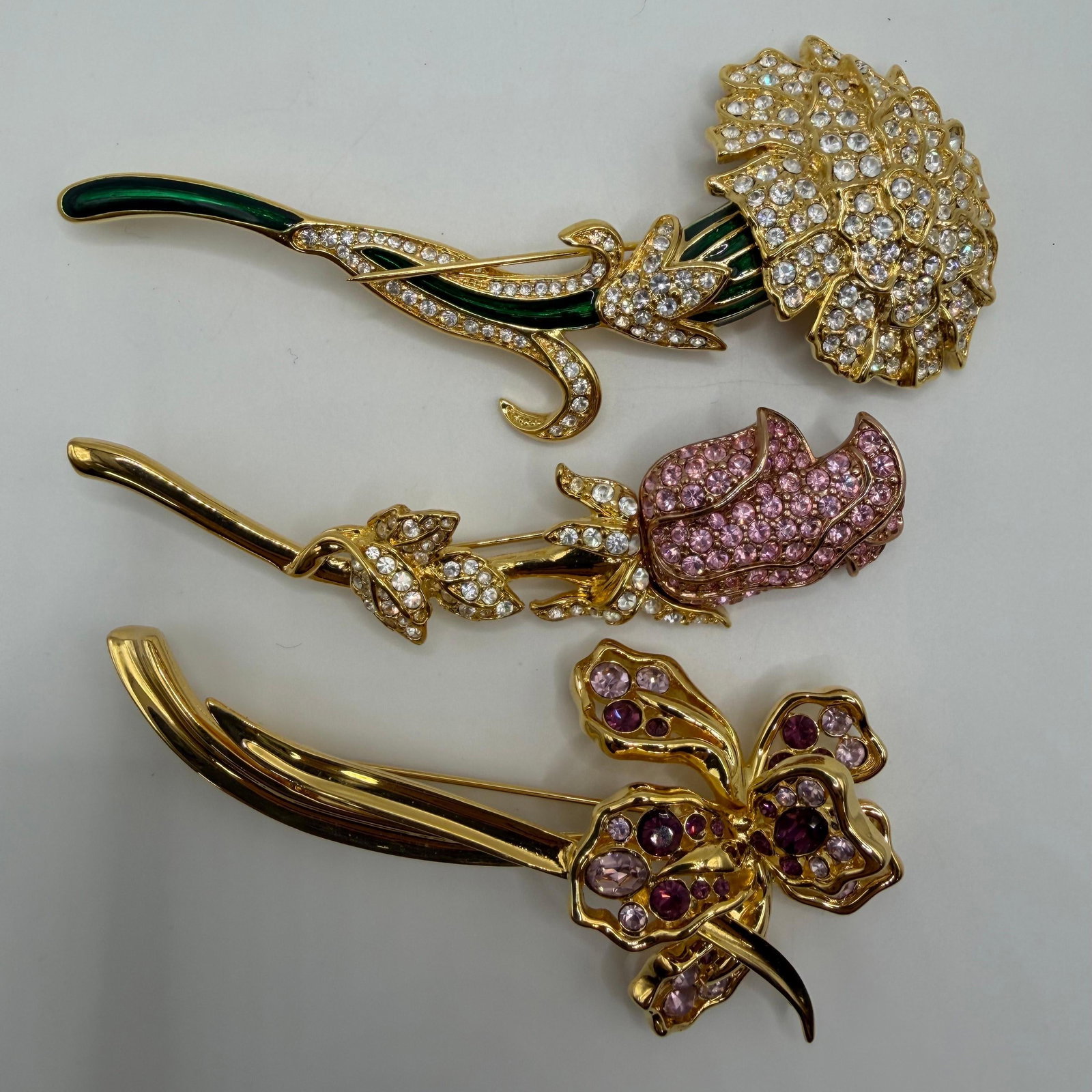 Three Nolan Miller Gold-Tone Floral Crystal Brooches Large Statement Pieces AB15: Collection of three signed Nolan Miller gold-tone floral brooches. Includes a large white crystal and green enamel flower brooch, a pink pave crystal rose brooch accented with clear rhinestones, 