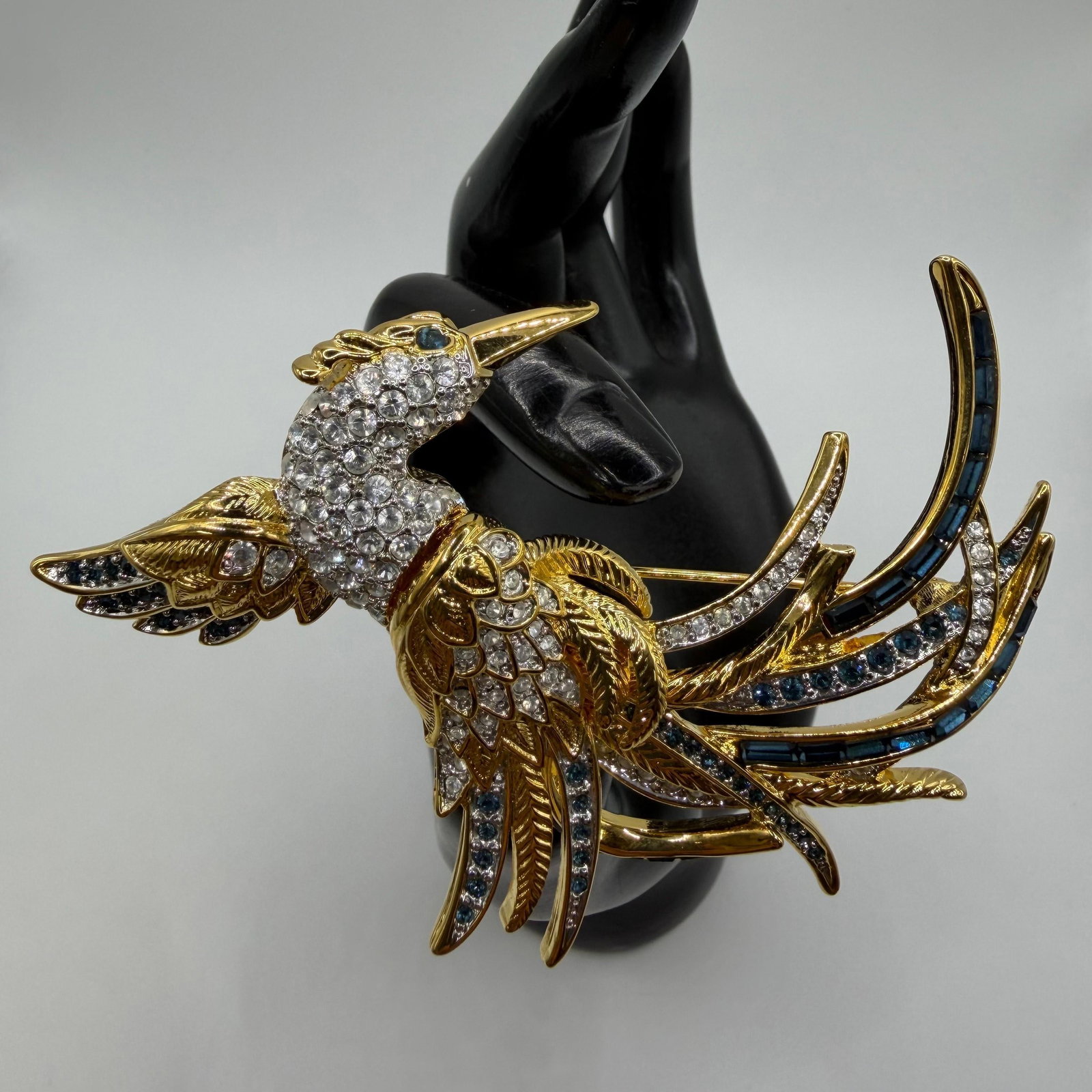 Nolan Miller Crystal and Blue Rhinestone Bird of Paradise Brooch Glamour Collection: Marked Nolan Miller. Large gold-tone Bird of Paradise brooch featuring a pave clear crystal body with blue sapphire-colored rhinestone accents, textured feather detailing, and dramatic sweeping t