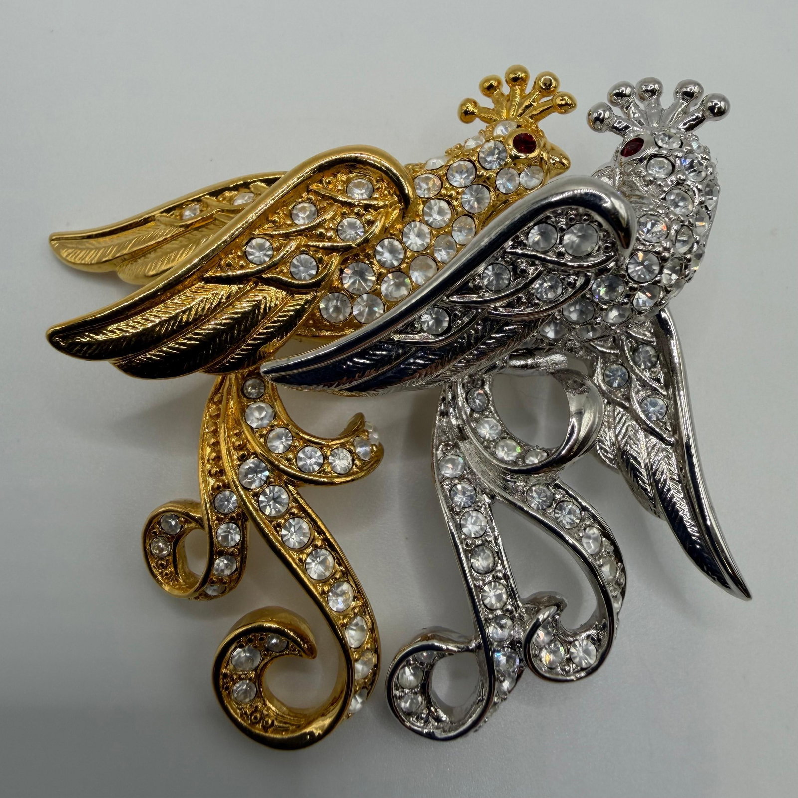 Nolan Miller Two Tone Crystal Lovebirds Designer Brooch Red Rhinestone: Marked Nolan Miller. Elegant two-tone gold and silver-tone lovebirds brooch adorned with clear faceted crystals and ruby-red crystal eyes. Features detailed feather engraving and graceful scrolling ta
