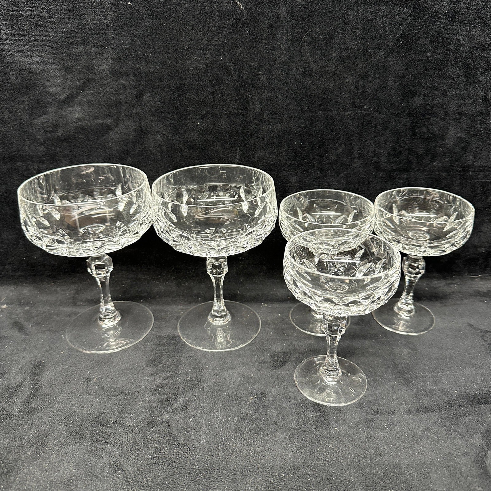 Group Of Five Cut Crystal Champagne Coupe Glasses: Group of five cut crystal champagne coupe glasses including two larger examples and three smaller examples. Features faceted stems and geometric cut decoration throughout. Light surface wear consisten