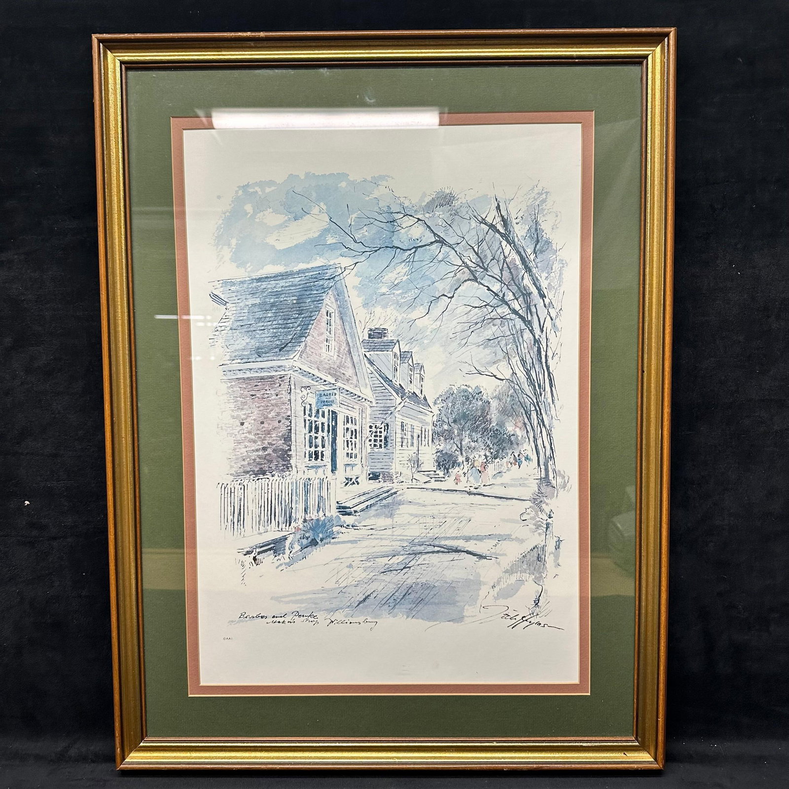 Framed Architectural Street Scene Print Williamsburg John Haymson Barber and Peruke Maker's Shop: Framed architectural street scene depicting historic buildings and figures along a tree-lined road, identified in the plate text as Williamsburg. Print signed in the lower right margin; additional han