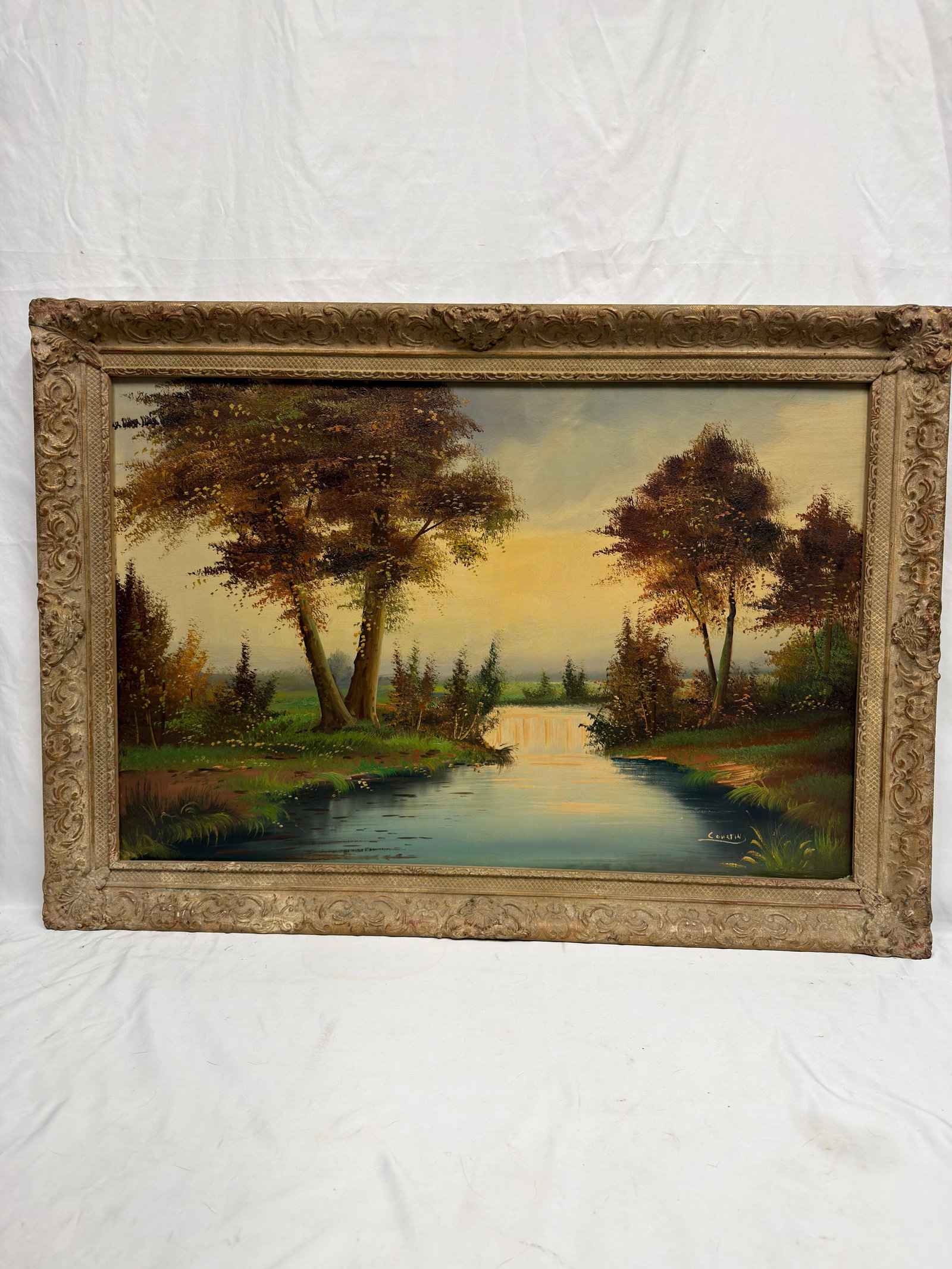 Signed Tranquil River Landscape Oil on Canvas Framed: Original oil painting on canvas depicting a peaceful river landscape bordered by mature trees and lush greenery beneath a softly illuminated sky. Signed by an unknown artist at the lower right. E