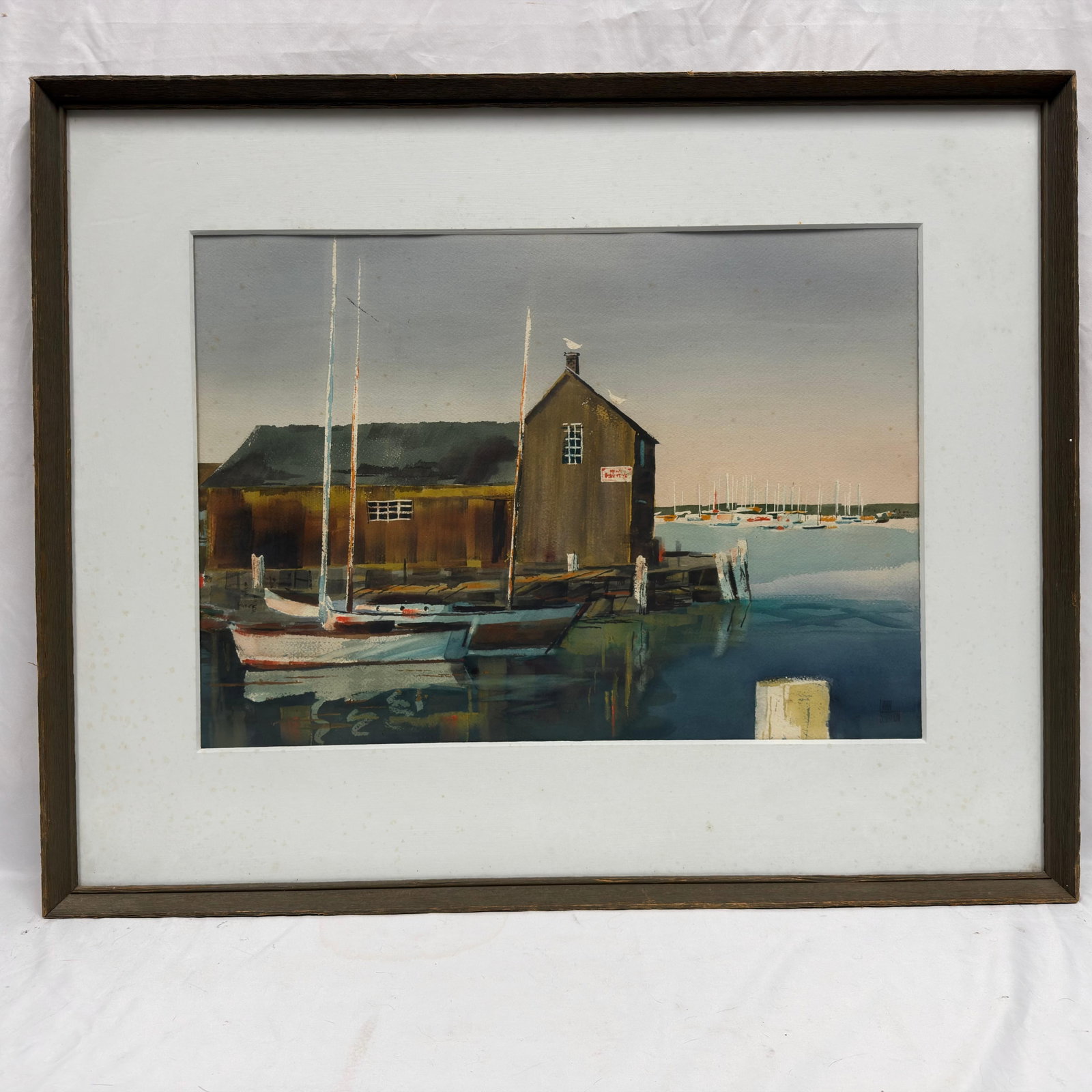 Larry Stratton Framed Harbor Landscape Art Print Sailboats at Marina: Framed decorative harbor scene print by Larry Stratton, depicting sailboats moored beside a weathered waterfront building with a marina in the distance. Rendered in muted coastal tones with a wide whi
