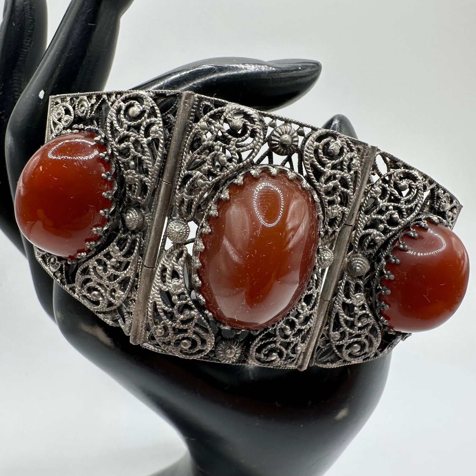 Filigree Panel Bracelet with Faux Red Cabochon Oval Setting Ornate Motifs: Wide panel bracelet with ornate filigree metalwork set with three large red cabochon stones. Hinged segments with box clasp closure. Unmarked and base metal non-silver; stones likely glass or faux sim