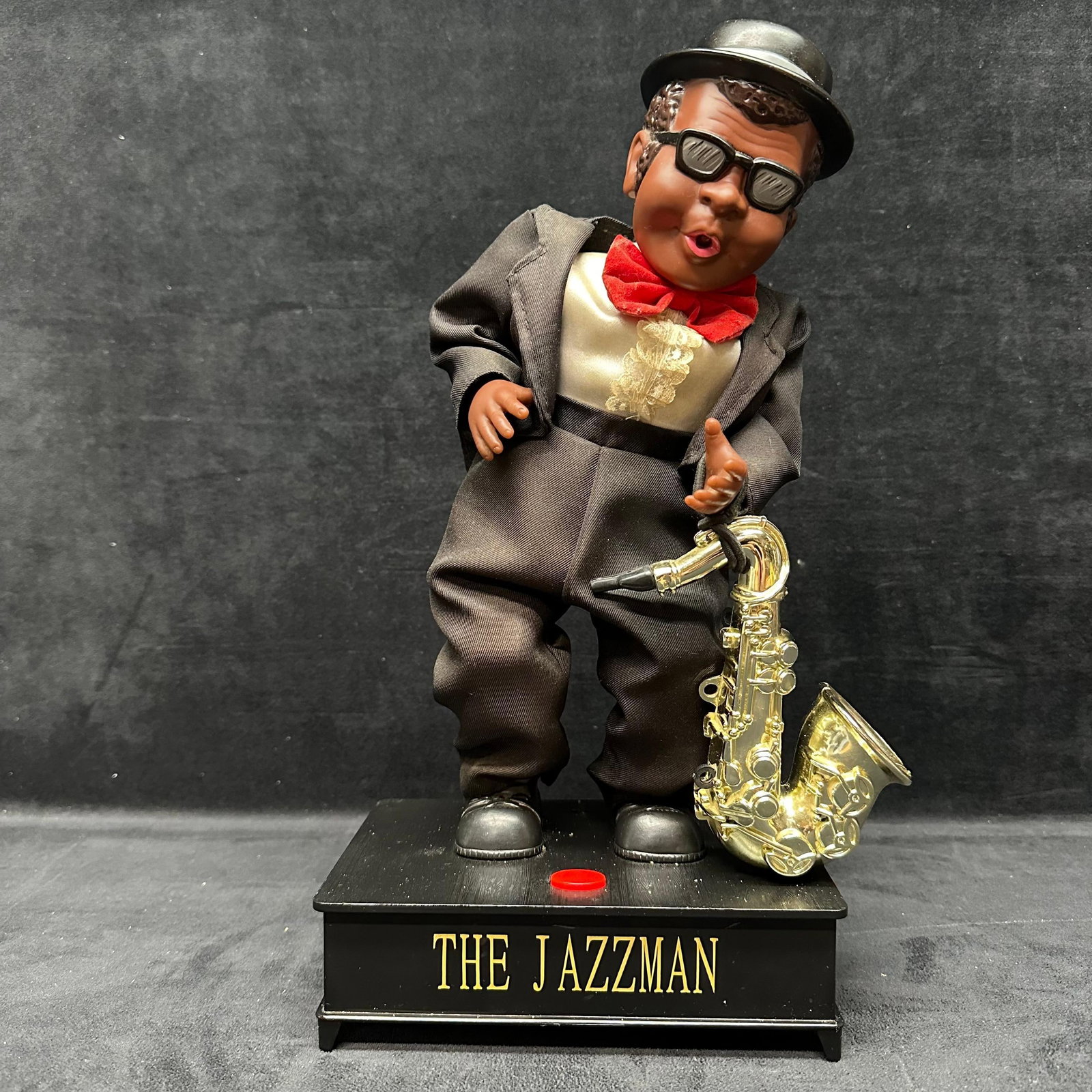 The Jazzman Singing Dancing Animated Sound Battery Operated Saxophone Player: Animated novelty figure titled The Jazzman depicting a saxophone player wearing a suit, hat, and sunglasses. Plastic figure with cloth clothing and metal-tone saxophone mounted on a black base.
