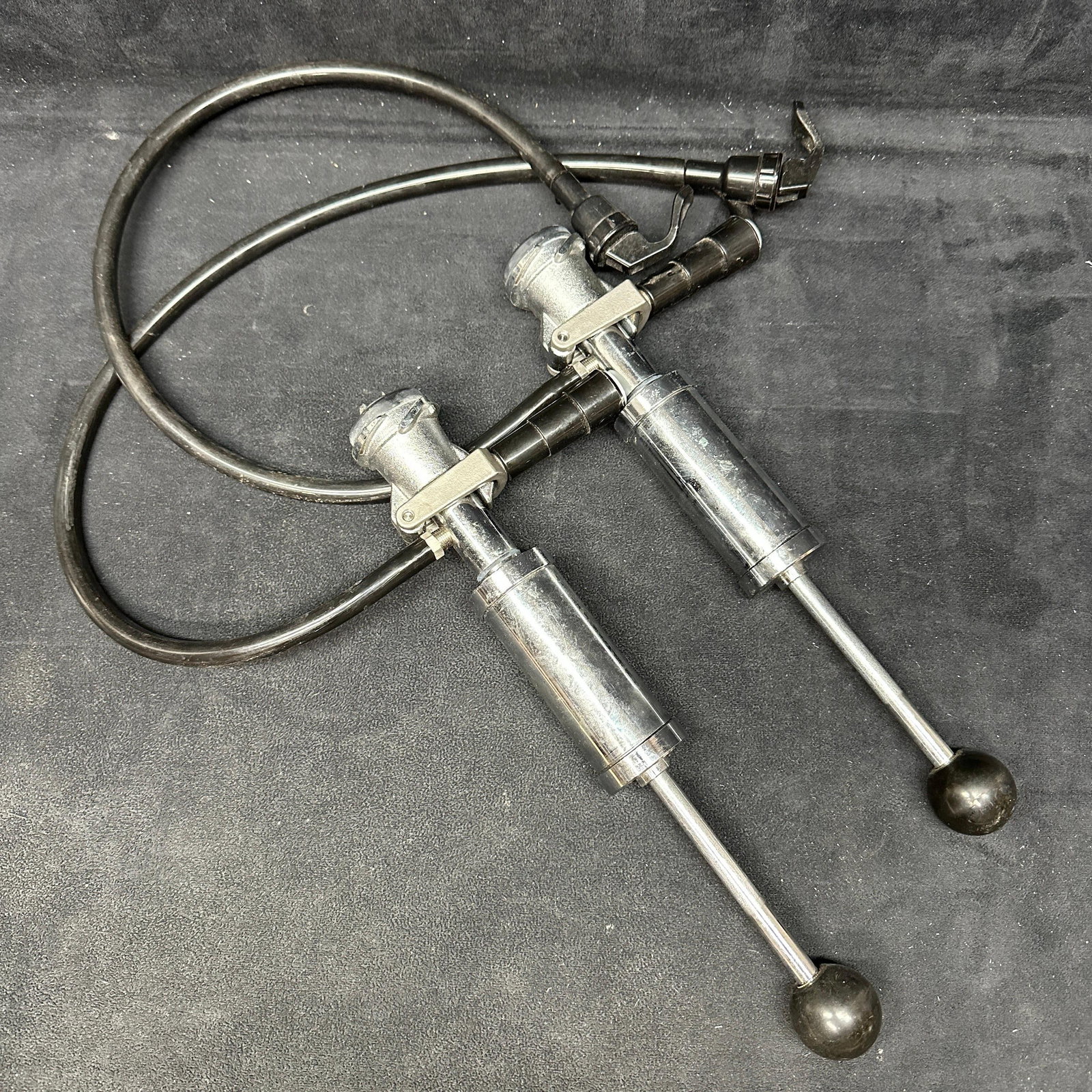 Pair of Stainless Steel Beer Keg Tap Pump Dispensers with Hoses: Set of two manual beer keg tap pump dispensers with metal pump bodies, ball handle plungers, and attached hoses. Each unit features a lever-style tap spout and standard keg coupler fittings. Showing