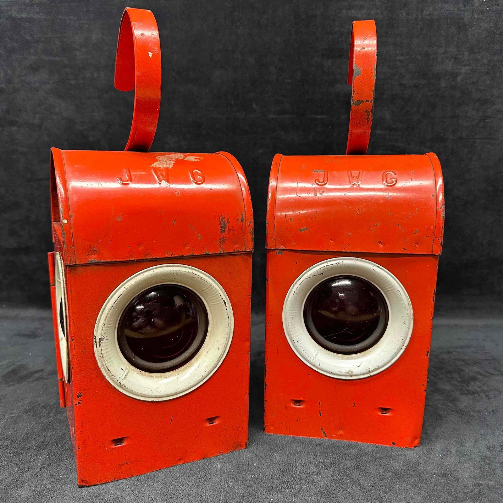 Pair of Vintage JWG Railway Industrial Red Metal Signal Lanterns: Pair of vintage metal signal lanterns, marked "JWG" on top. Each lantern is constructed of heavy red-painted metal with curved hanging hooks and fitted with round glass lenses framed in white.