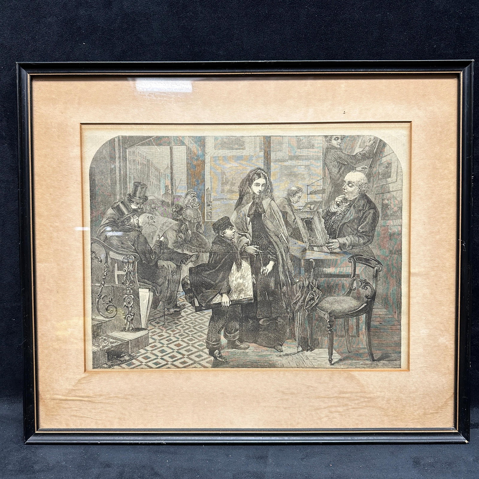 Victorian Style Print Shop Interior Genre Scene Framed Engraving Art Print: Black and white engraving depicting a Victorian-style era print or publishing shop interior with figures examining prints and reading materials. Includes seated and standing figures in period dress wi