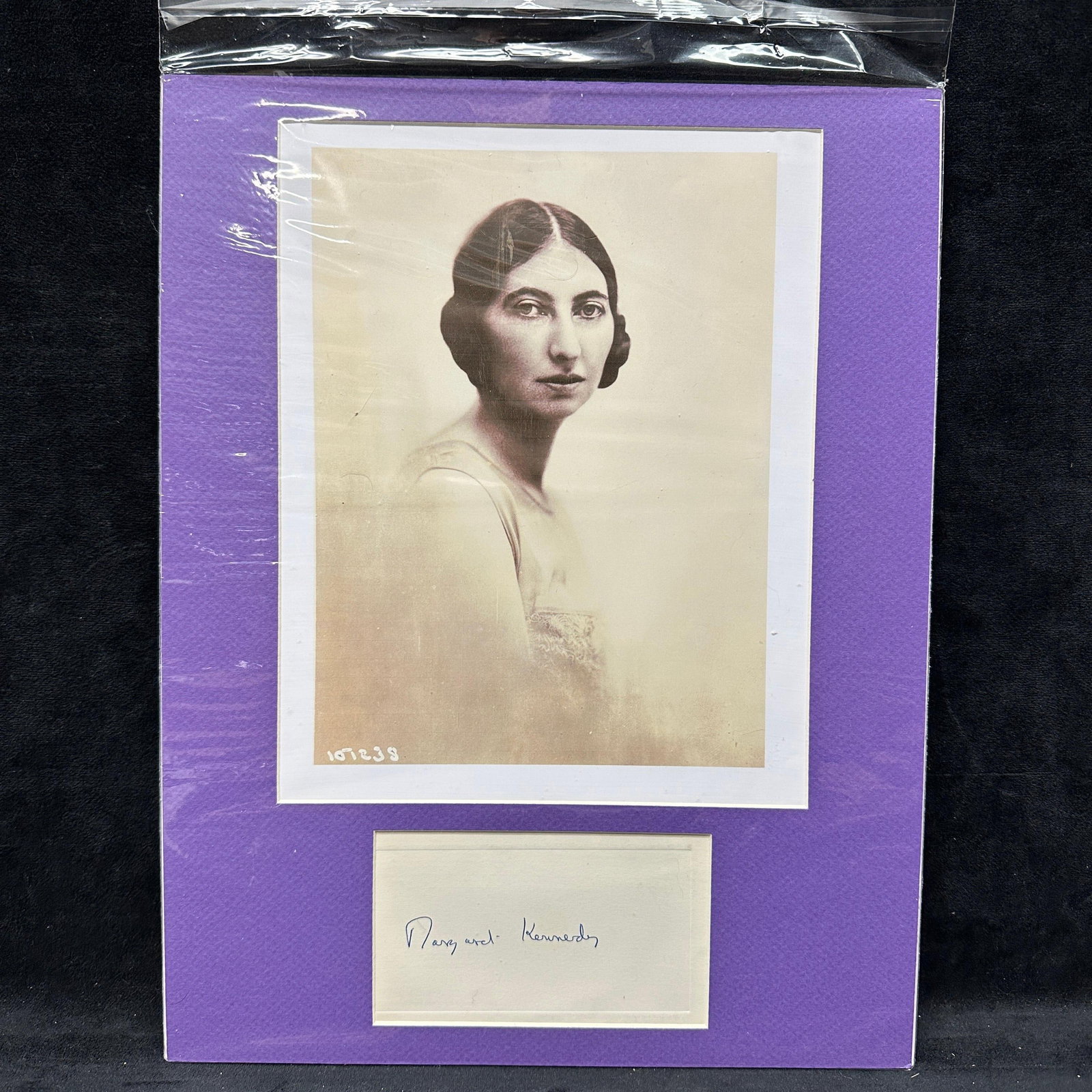 Margaret Kennedy Signed Autograph Display With Portrait: Framed autograph display featuring a clipped handwritten signature of Margaret Kennedy mounted beneath a vintage black and white portrait photograph print. Ink signature on paper, presented in a purpl