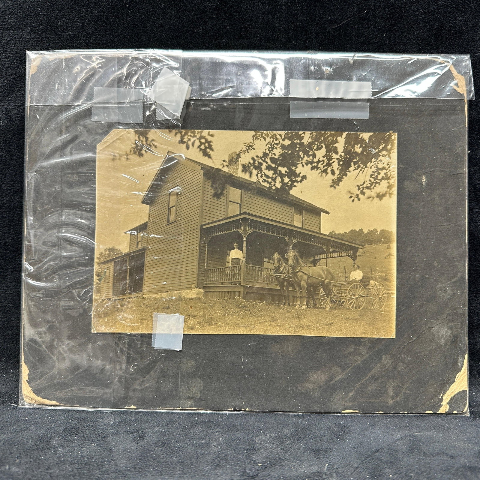 Antique Rural Farmhouse Photograph With Horse Drawn Wagon: Antique sepia tone photograph depicting a rural farmhouse scene with two figures, horse drawn wagon, and pair of horses in front of a wood frame home with wraparound porch. Mounted on black backing