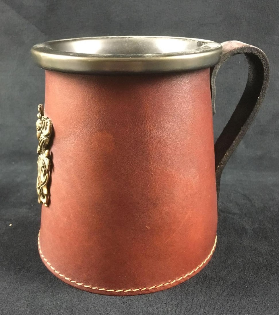 English Beer Mug with Leather Sleeve: English Beer Mug with Leather Sleeve. This mug is in great condition. The insert is made of ceramic. The exterior of the leather sleeve has a metal coat of arm decoration attached to it. Mug measures 
