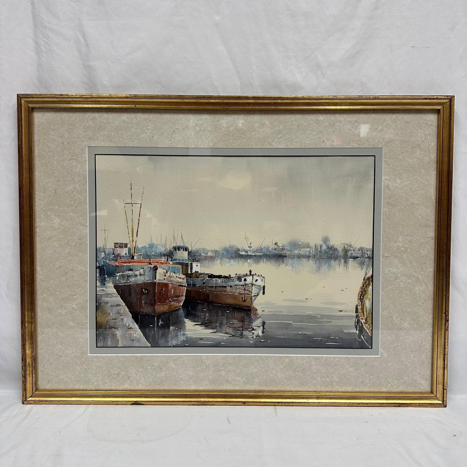Watercolor Harbor Scene with Docked Boats Signed Framed Seascape: Framed watercolor painting depicting two docked boats along a calm harbor with distant shoreline and soft atmospheric light. Signed lower right by the artist, unknown. Housed in a gold-tone frame with