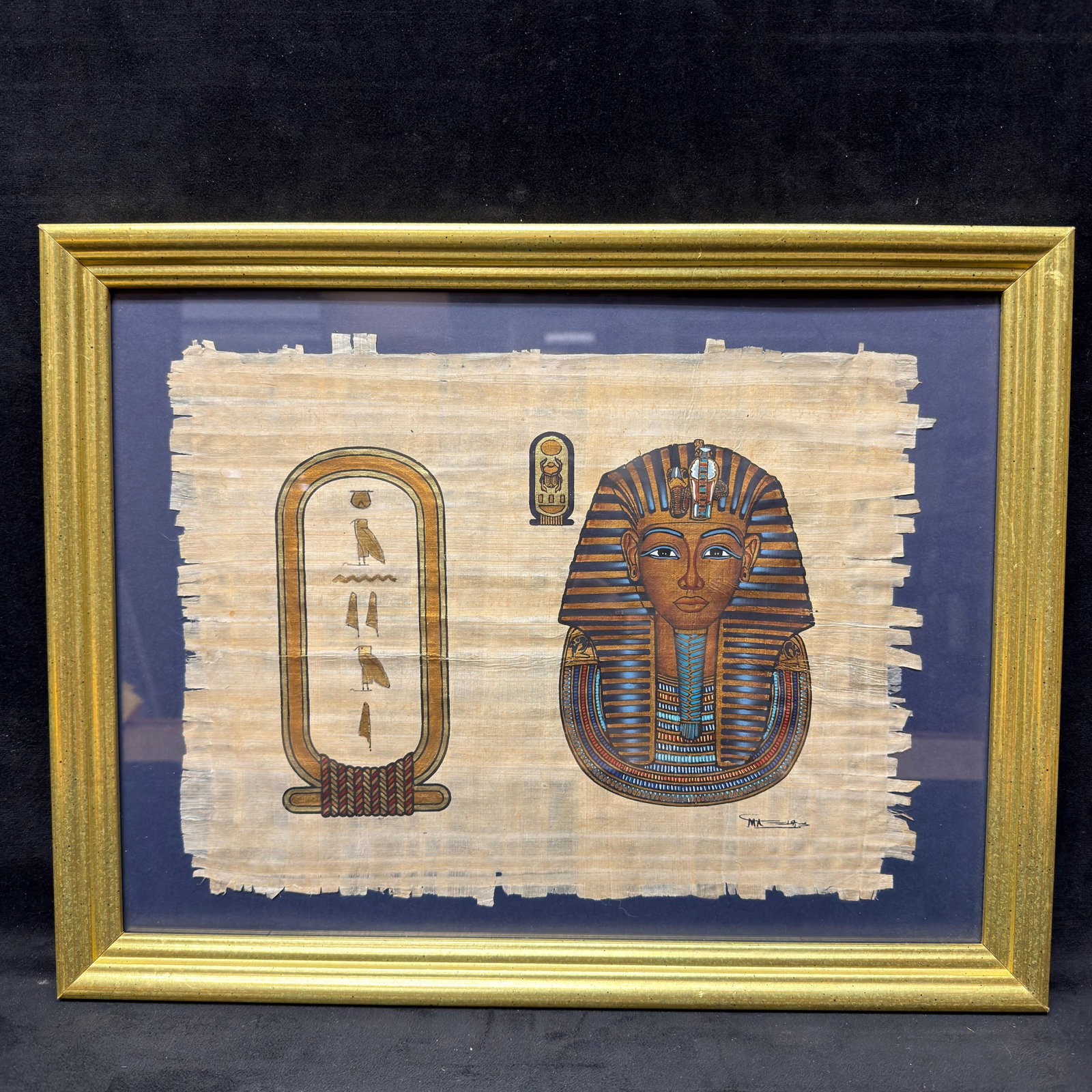 Egyptian Pharaoh Tutankhamun Folk Art Painting Signed: Egyptian-style painting on papyrus-style woven paper depicting the funerary mask of Pharaoh Tutankhamun alongside a hieroglyphic cartouche. Executed in black, blue, red, and metallic gold tones on pap