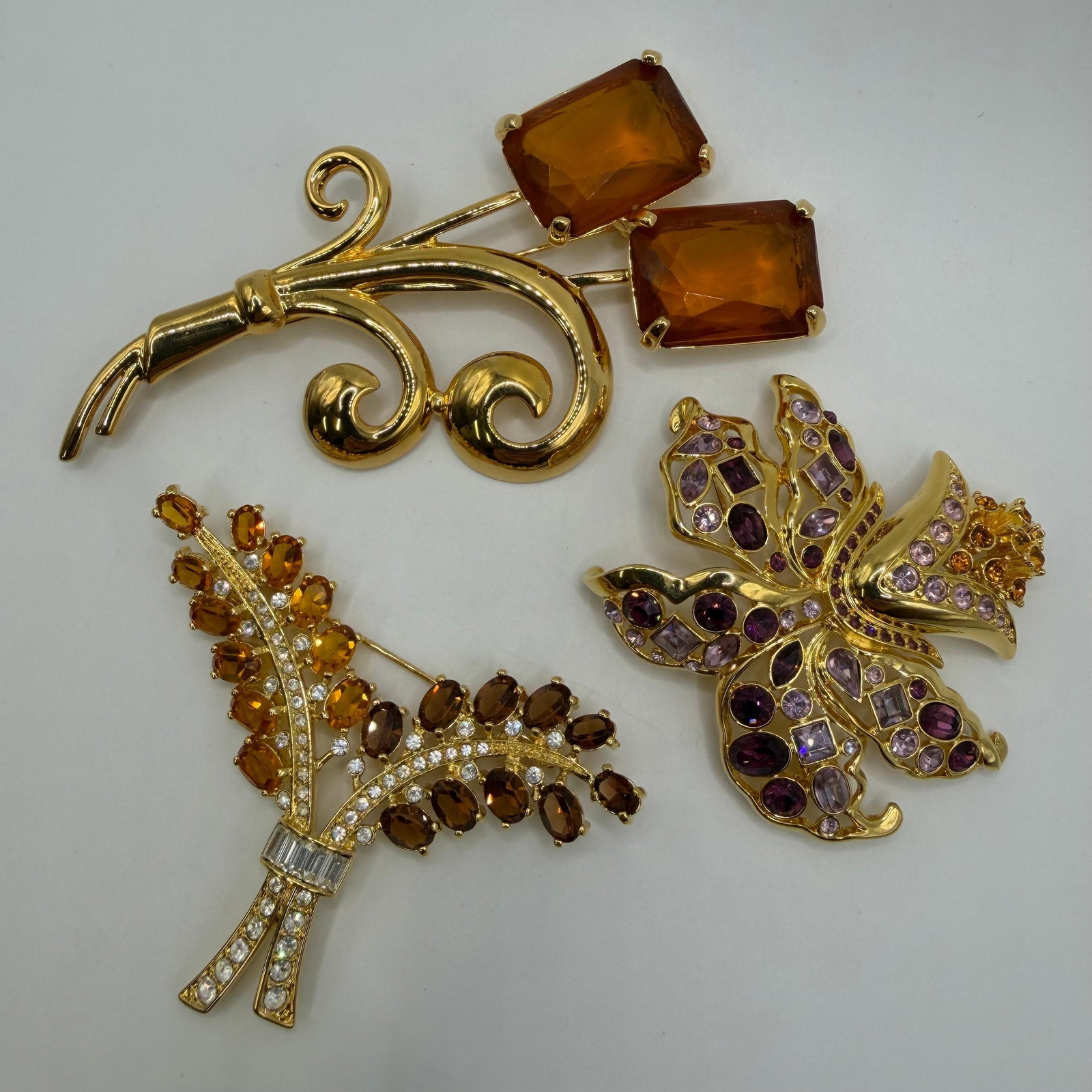 Three Gold Tone Nolan Miller Designer Rhinestone Brooches Amber Purple Orange: Lot of three Nolan Miller designer brooches in gold-tone finish. Includes an abstract scroll brooch with large amber-colored faceted crystals, a floral spray brooch set with amber and smoky topaz-colo