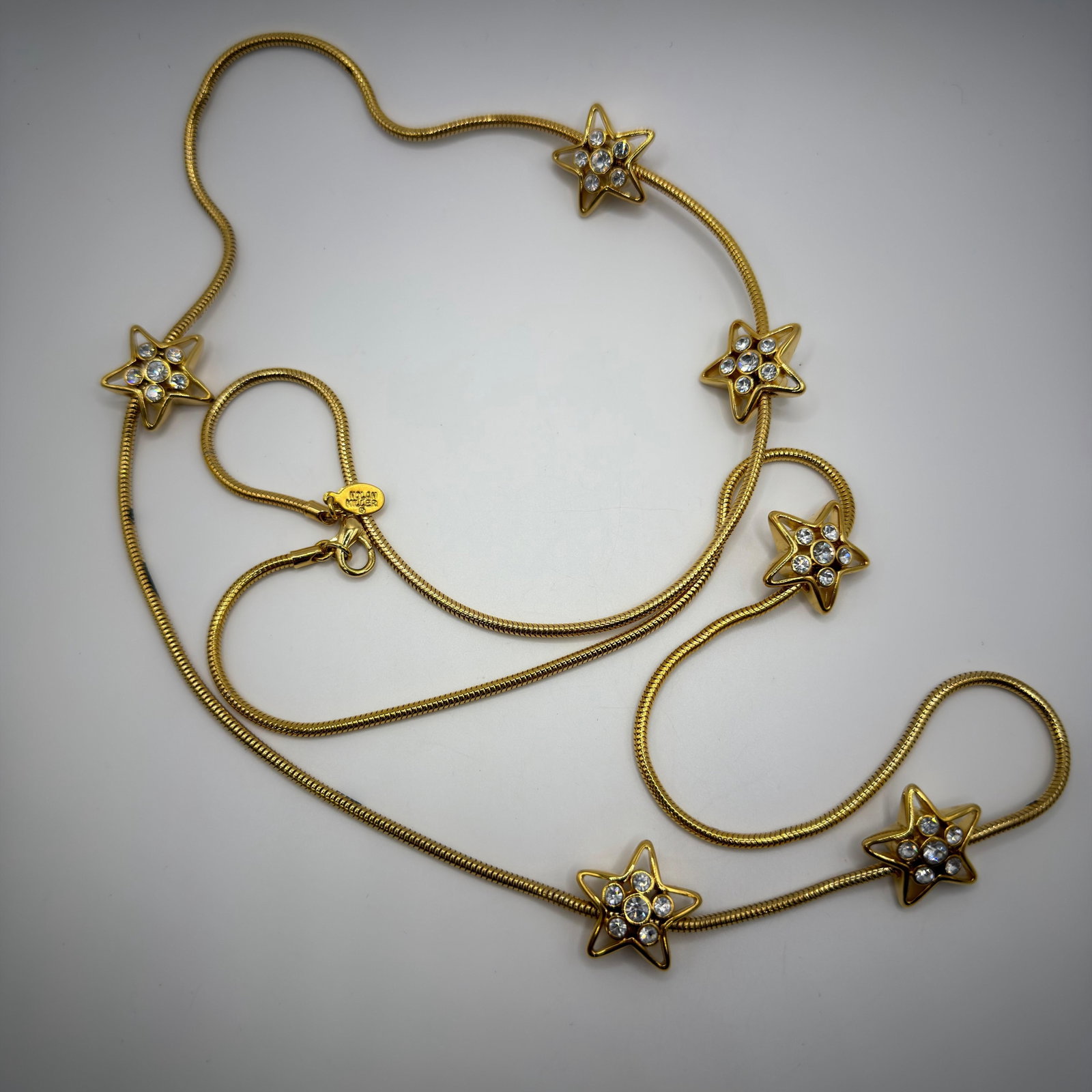 Nolan Miller Crystal Rhinestone Star Snake Chain Necklace Long: Nolan Miller gold-tone snake chain necklace featuring six open star stations accented with clear round-cut crystals/rhinestones. Finished with a lobster clasp and Nolan Miller signature tag. This item