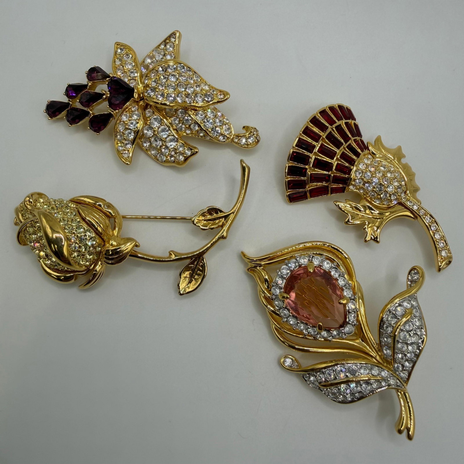 Group of Four Nolan Miller Floral Rhinestone Designer Brooches AB23: Collection of four signed Nolan Miller designer costume brooches featuring floral motifs. Includes a clear rhinestone flower with deep purple accent stones, a long-stem rose, a red enamel and rhinesto