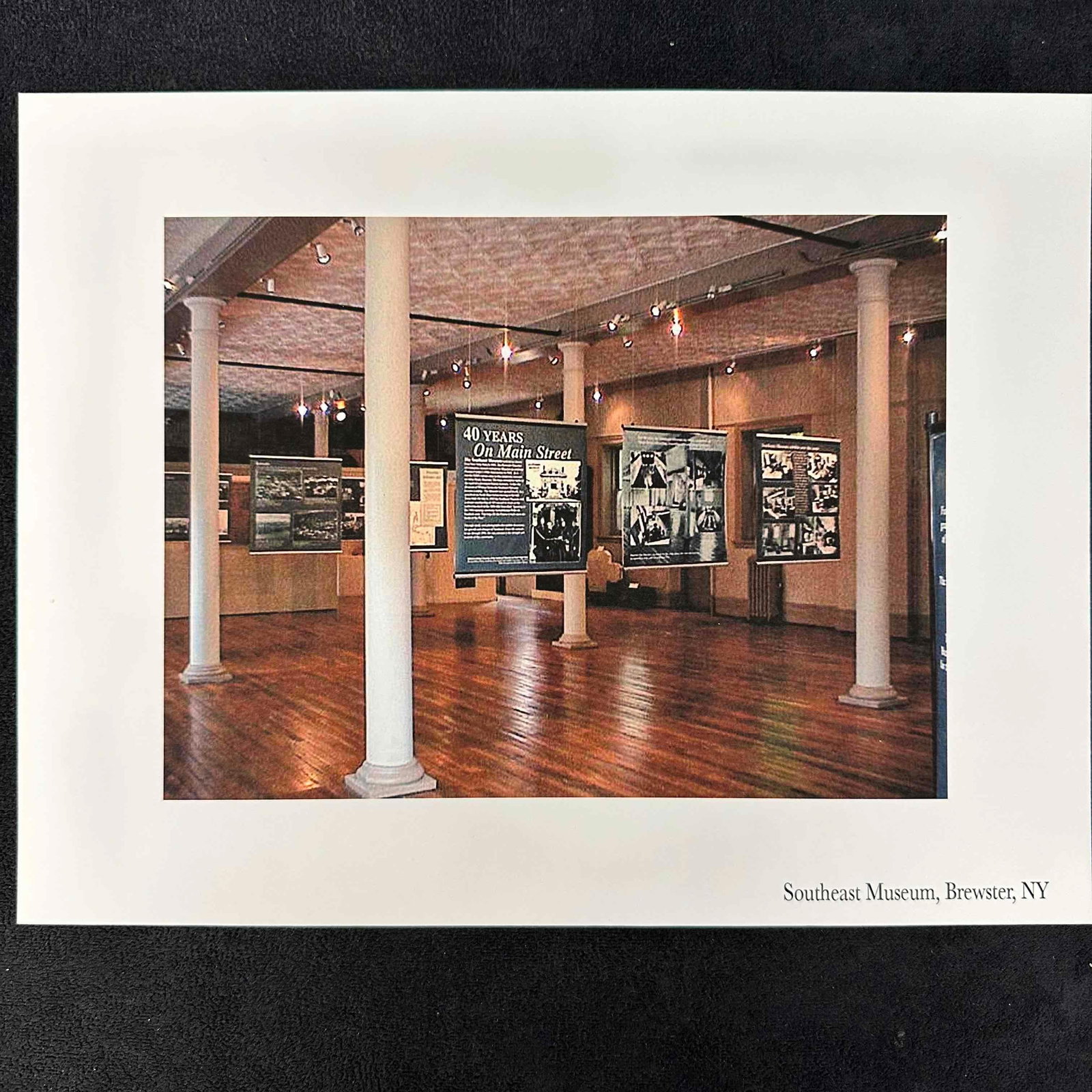 Southeast Museum Brewster NY Exhibit Photography Print: Color photographic print showcasing the Southeast Museum in Brewster, New York. The image captures the warm wooden floors, tall white columns, and hanging exhibit panels, including the featured displa