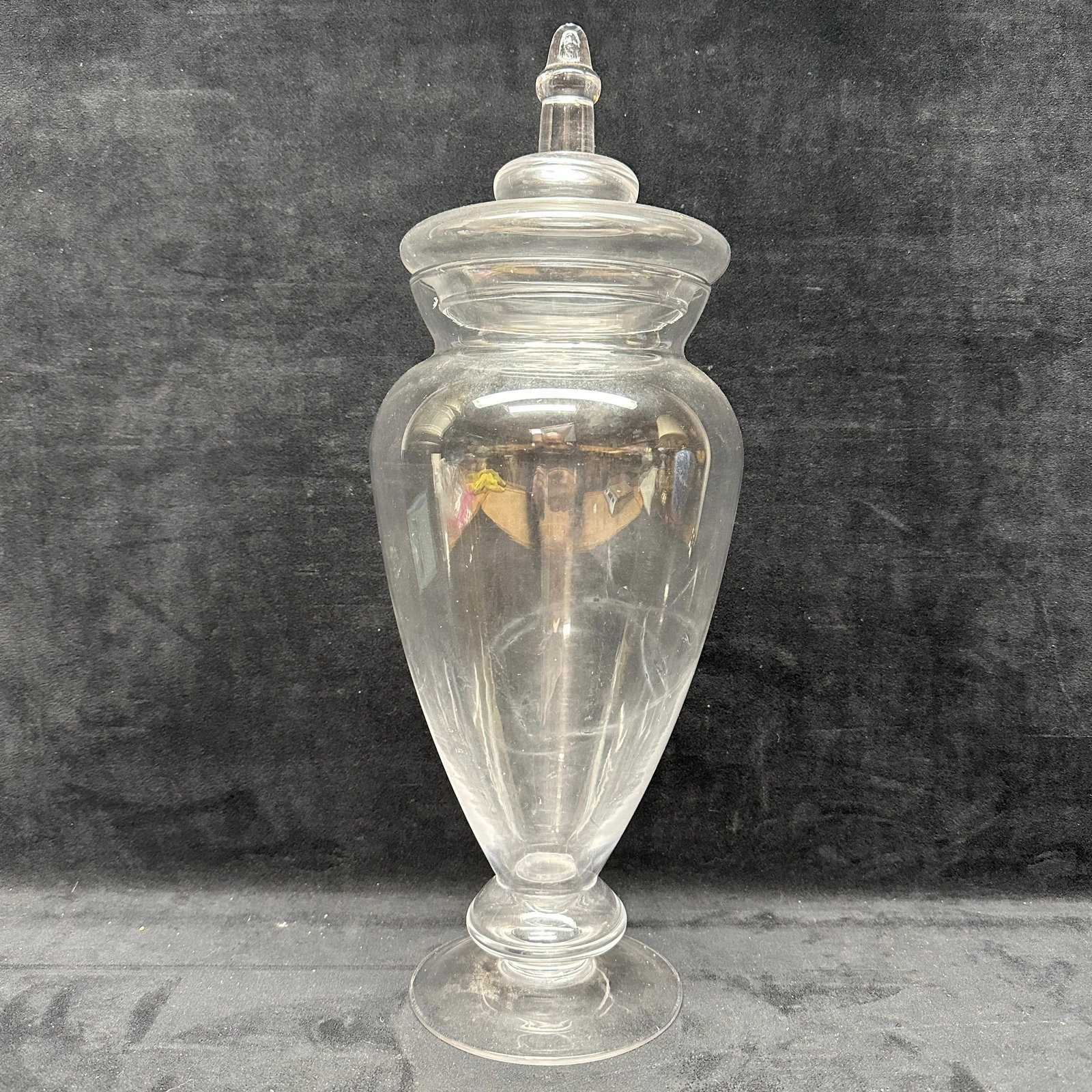 Clear Glass Apothecary Jar With Lid Tall Covered Candy Storage: Clear glass apothecary-style jar with fitted lid and finial top. Tall form with flared footed base and wide mouth opening. Unmarked. Shows light surface wear and minor scratches consistent with age an