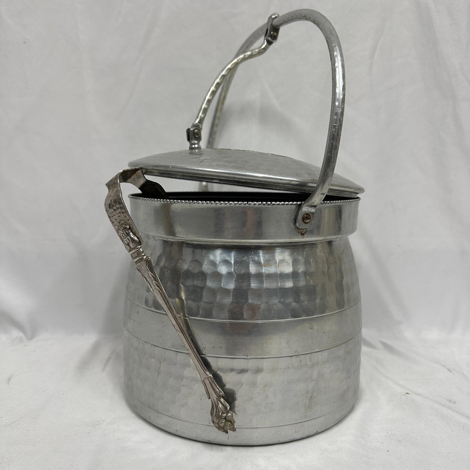 Vintage Hammered Aluminum Ice Bucket With Lid And Tongs: Vintage hammered aluminum ice bucket with fitted lid and swing handle, featuring decorative embossed fruit and leaf motif on lid. Includes a pair of metal serving tongs. Unmarked. Shows surface wear, 