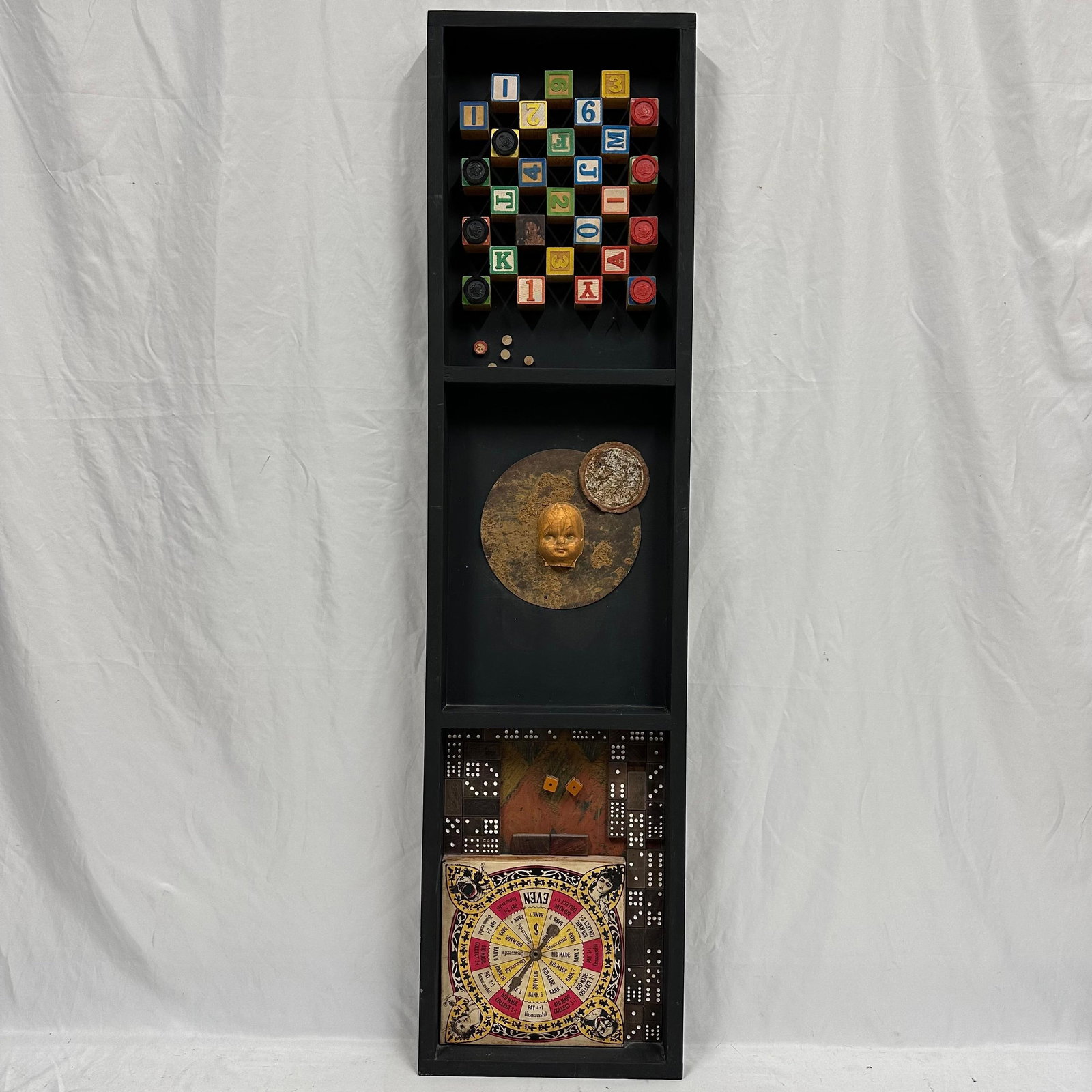 Tall Mixed Media Shadow Box Wall Art Featuring Board Game Pieces and Elements Doll Face: Large vertical mixed media wall art presented in a deep black shadow box frame. The composition features multiple assemblage sections, including vintage-style wooden letter and number blocks, dominoes