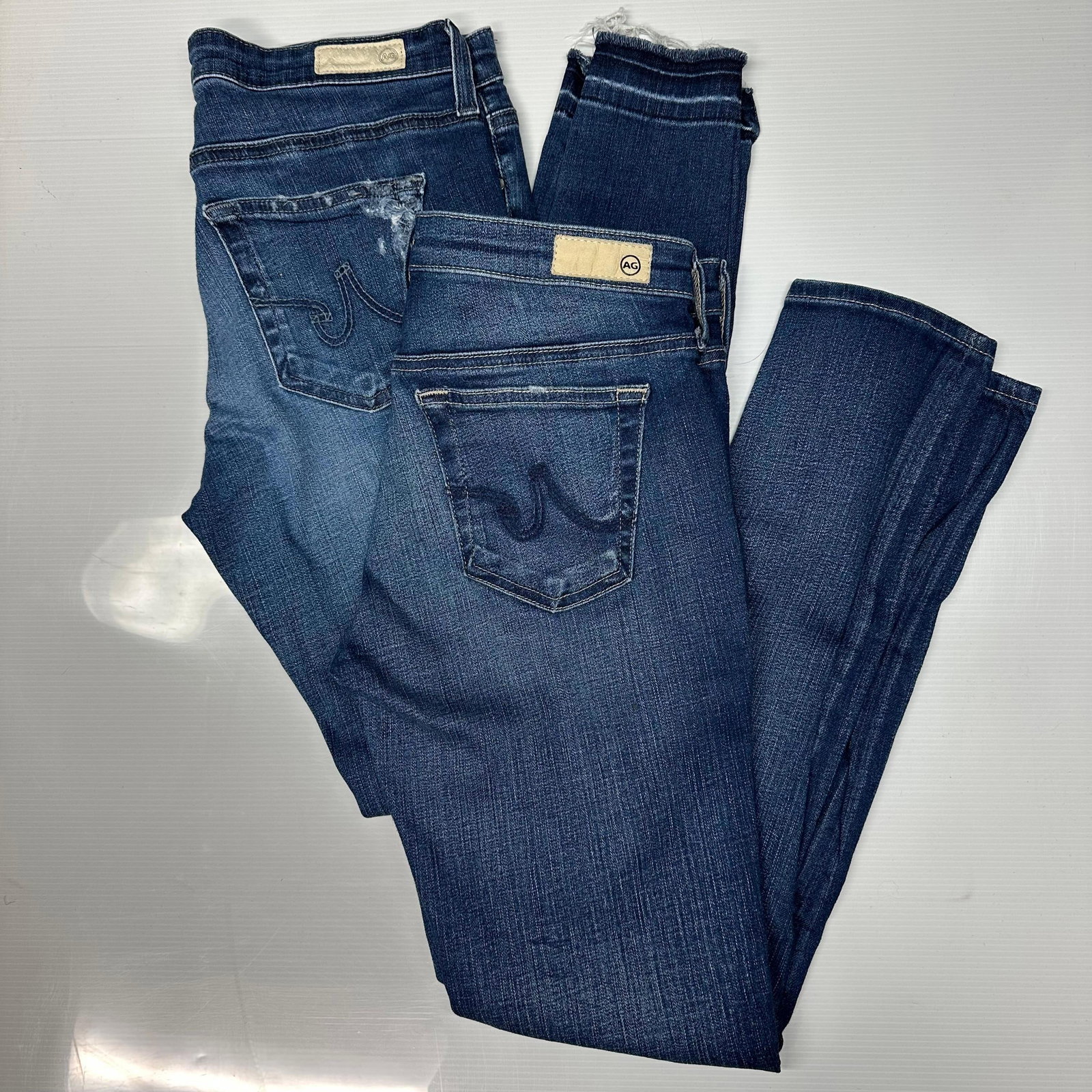 Lot of 2 AG Adriano Goldschmied Women Jeans Size 27R and 28R: The lot includes two pairs of women’s AG Adriano Goldschmied denim jeans. Both pairs appear to be AG The Legging Ankle style with slim fit and ankle-length legs. Medium blue wash with light fadi
