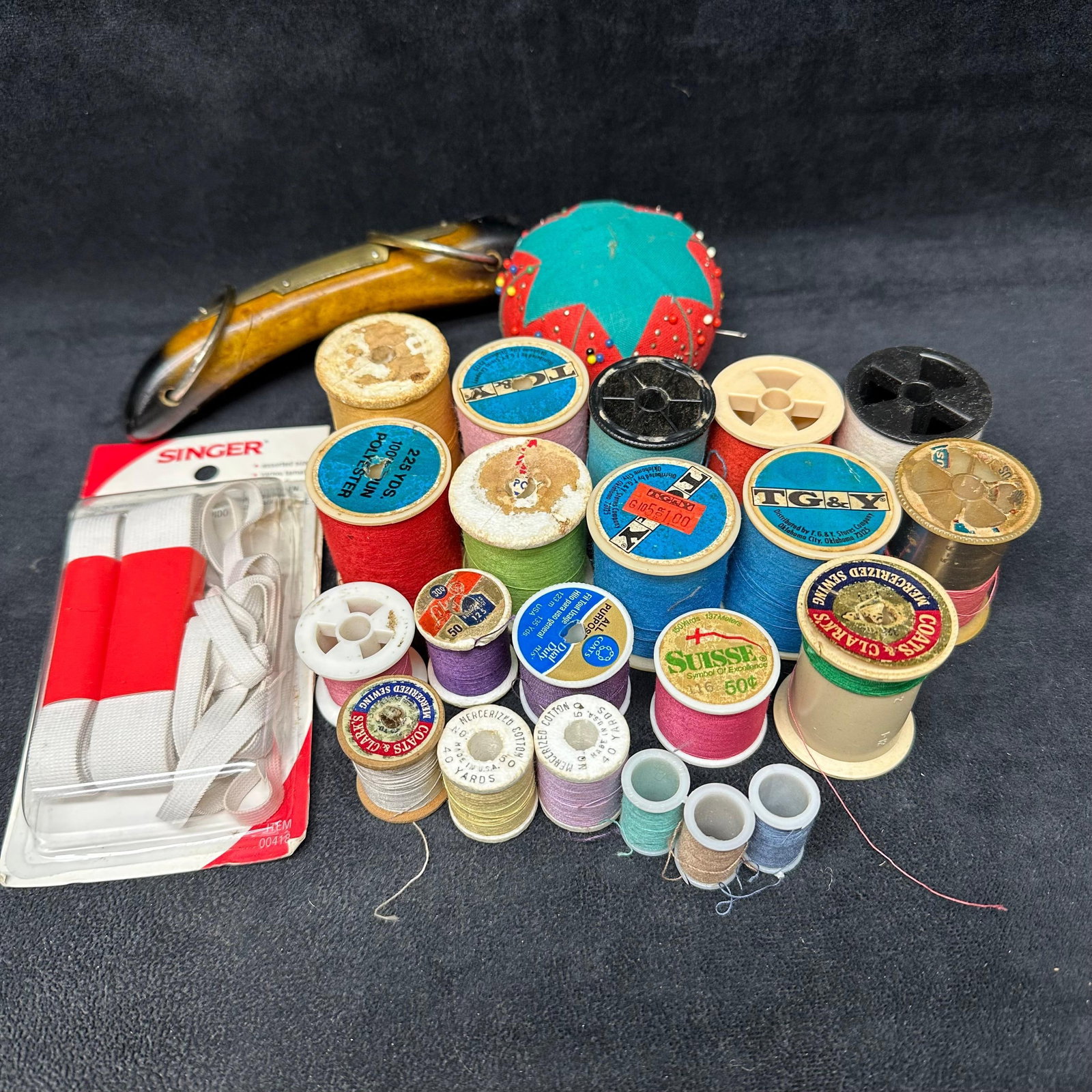 Vintage Sewing Notions Lot Thread Spools Pincushion Bodkin Singer Elastic: Mixed lot of vintage sewing notions, including assorted thread spools in various colors and brands, a pincushion with pins, a metal bodkin or sewing tool, and packaged Singer elastic. Spools show a va