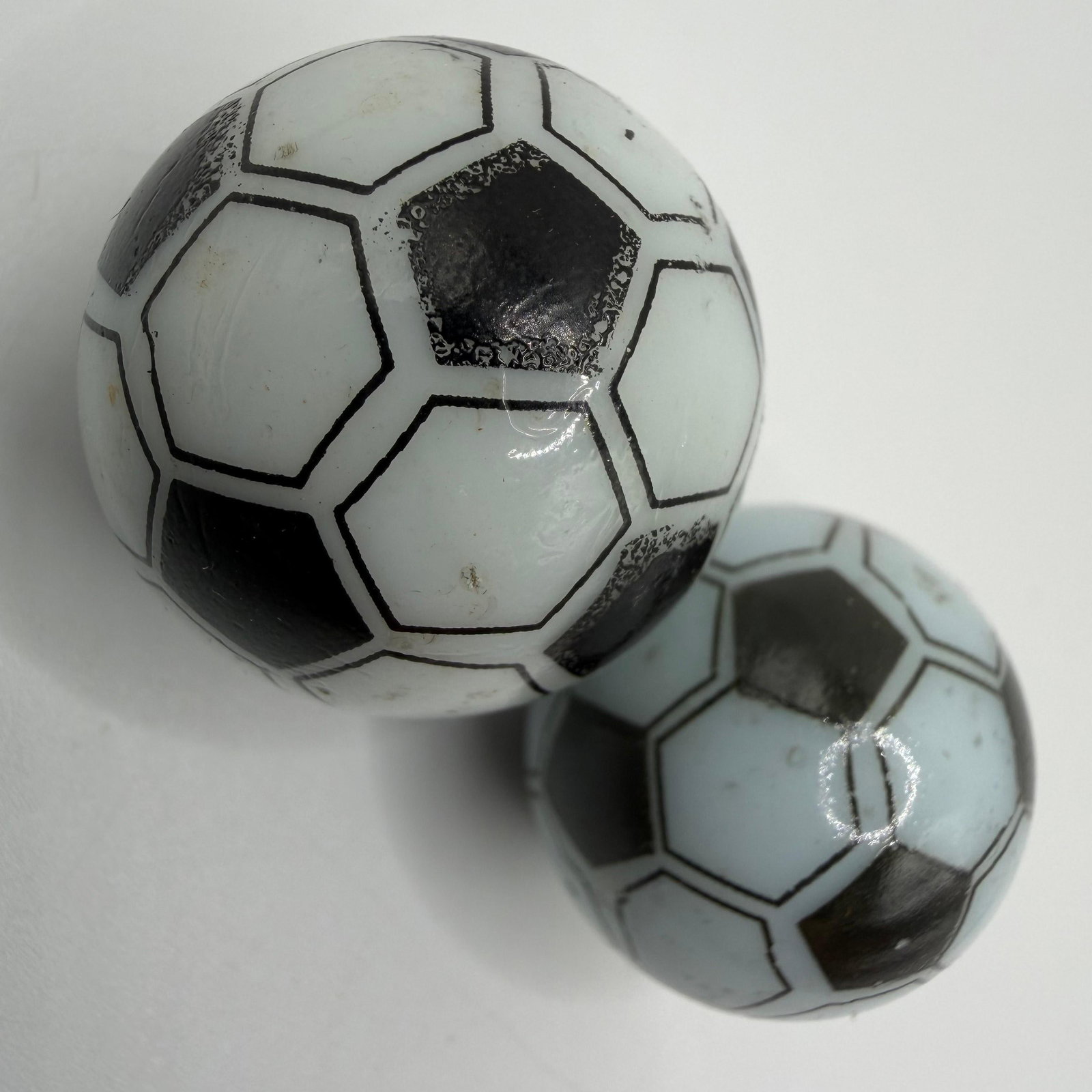 Two Soccer Ball Glass Shooter Marbles Black and White: Two 23mm and 25mm glass soccer ball marbles. Showing heavy play wear with surface scratches and markings throughout. These items are pre-owned, with noticeable wear, minor imperfections, and surf