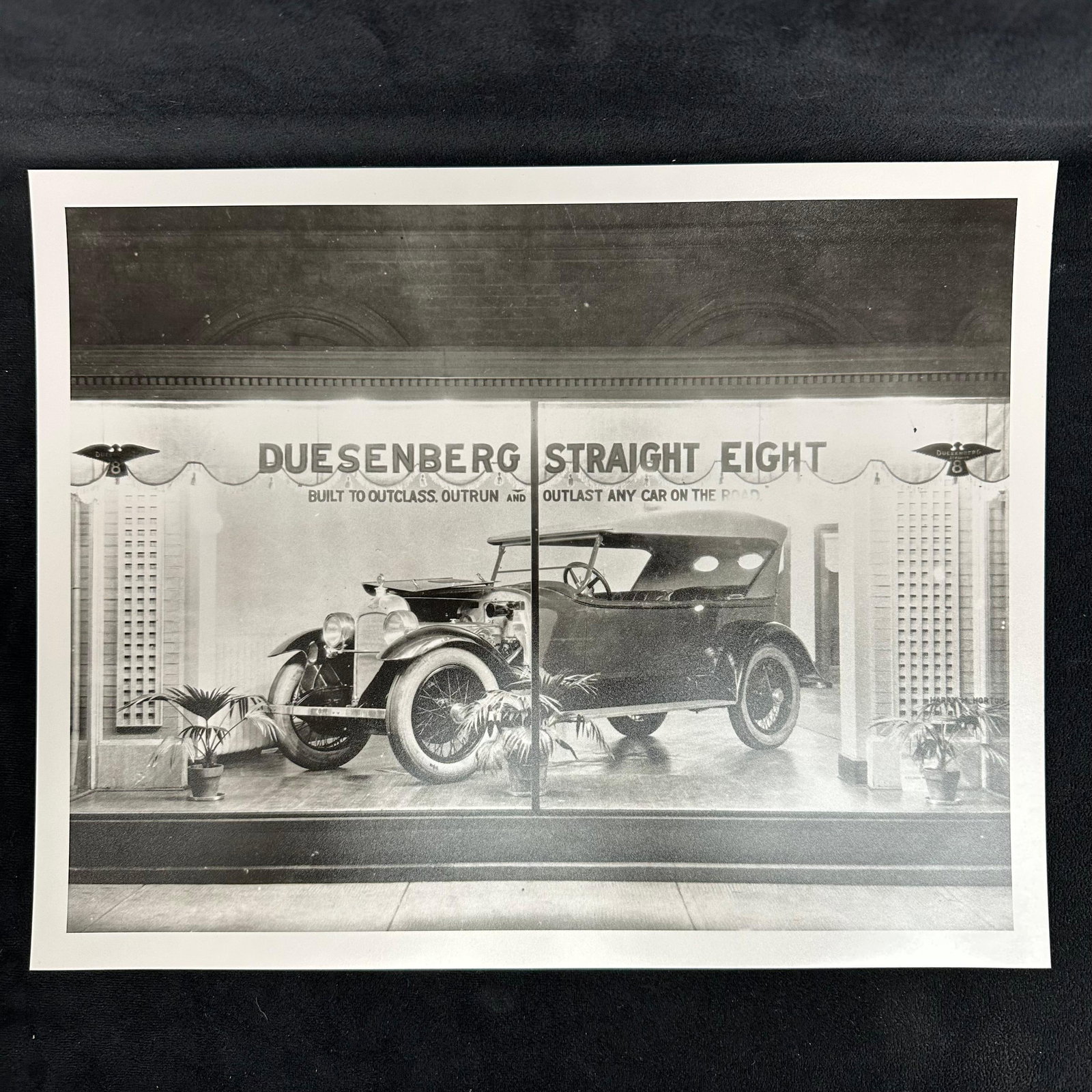 Duesenberg Straight Eight Automobile Display Analog Photo Print: Black and white analog photographic print depicting a showroom display of a Duesenberg Straight Eight automobile. The image captures the elegance of the early luxury car, showcased with signage