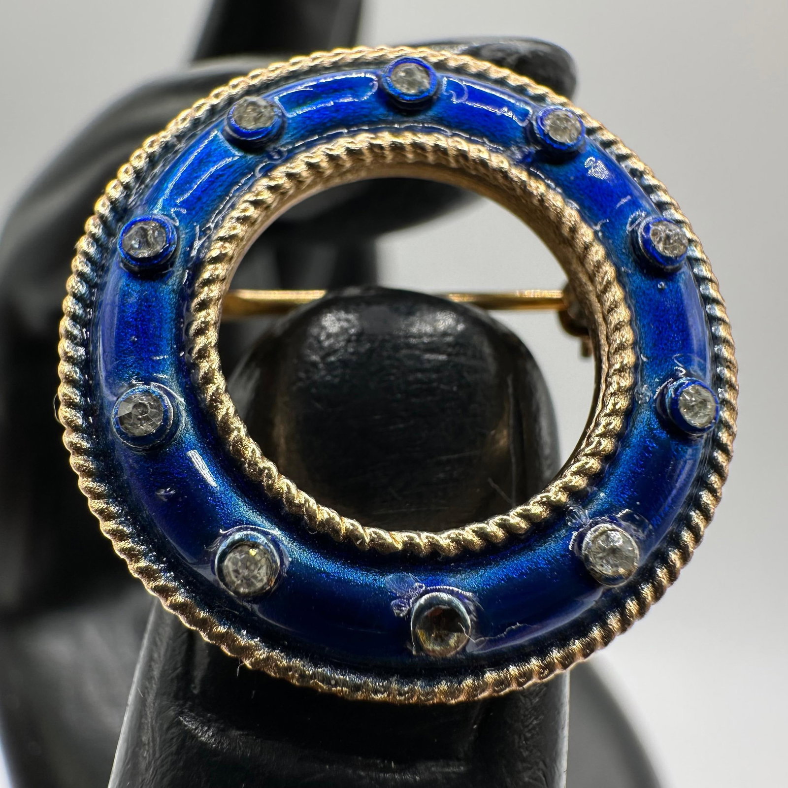 Crown Trifari Royal Blue Enameled Circle Rhinestone Accent Gold Tone Brooch: This vintage Trifari enameled circle brooch features a royal blue enamel finish with gold-toned roping and rhinestone accents. Vintage crown symbol Trifari hallmark on the back. One stone setting is m
