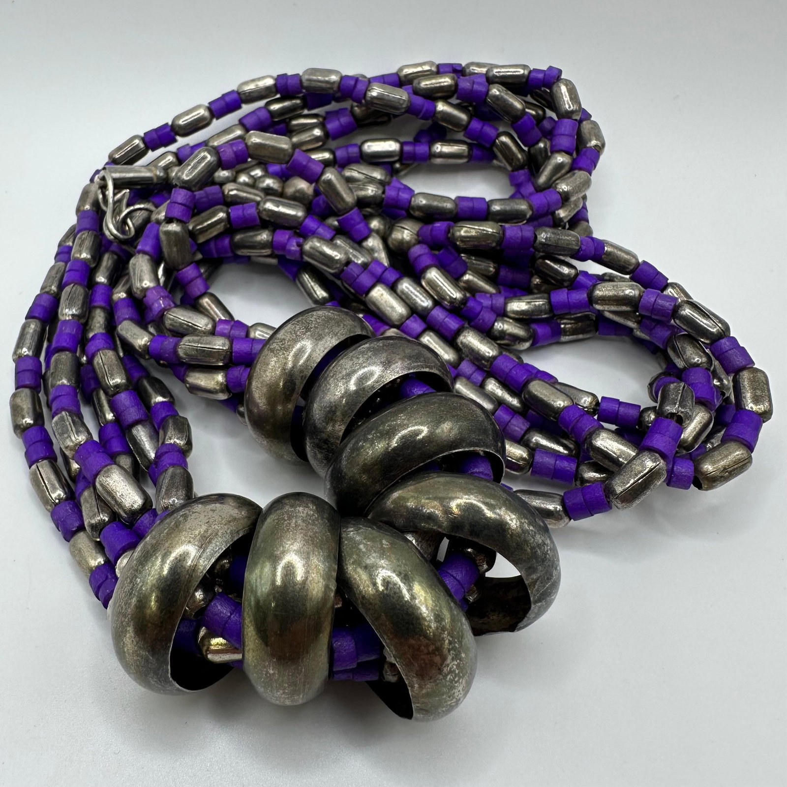 Multi Strand Beaded Necklace with Large Metal Ring Accents Purple Beads: Statement style multi-strand necklace featuring multiple strands of purple spacer beads and silver-tone barrel beads. The strands join at the front with a series of large oxidized silver-tone ring spa