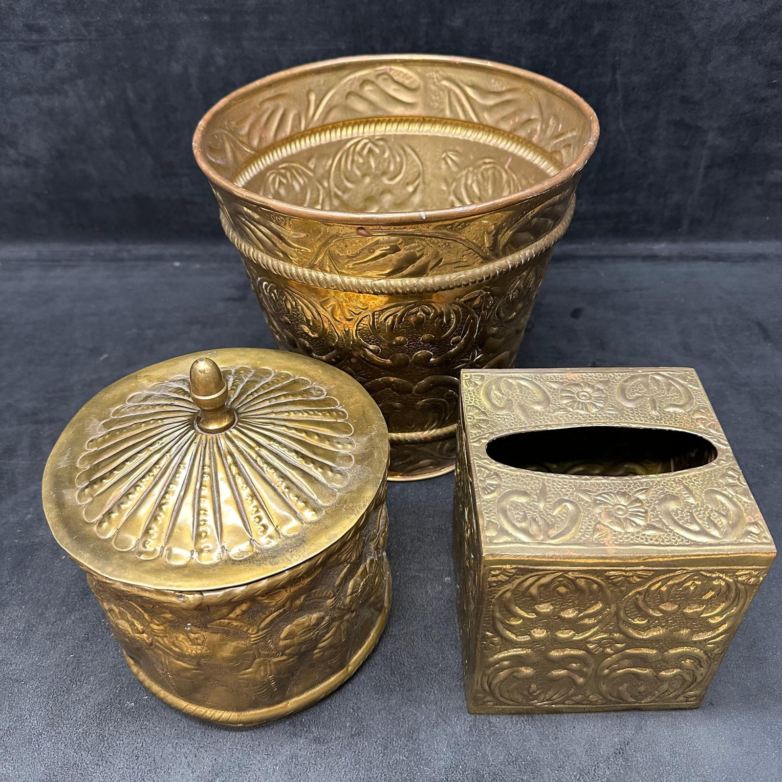 Embossed Brass Vanity Set Wastebasket Covered Jar and Tissue Box: A three-piece embossed brass vanity set including a wastebasket, a lidded jar, and a square tissue box cover. Each piece features ornate floral and scroll work designs with a warm, aged patina. Light 