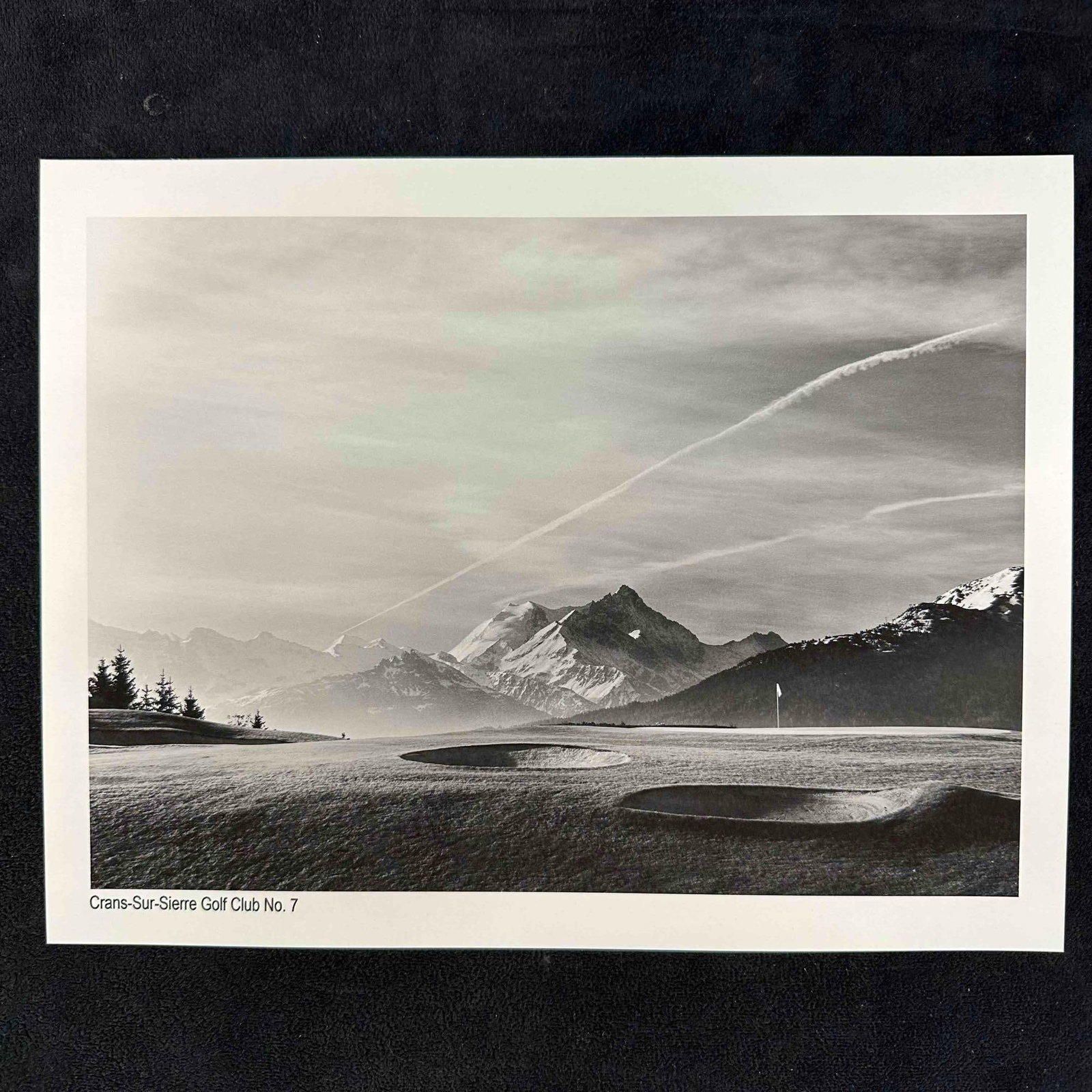 Crans-Sur-Sierre Golf Club No. 7 Black & White Landscape Photography Print: Black-and-white photograph featuring Crans-Sur-Sierre Golf Club’s 7th hole with dramatic mountain views in the background. The image beautifully captures the contrast between the rolling greens,