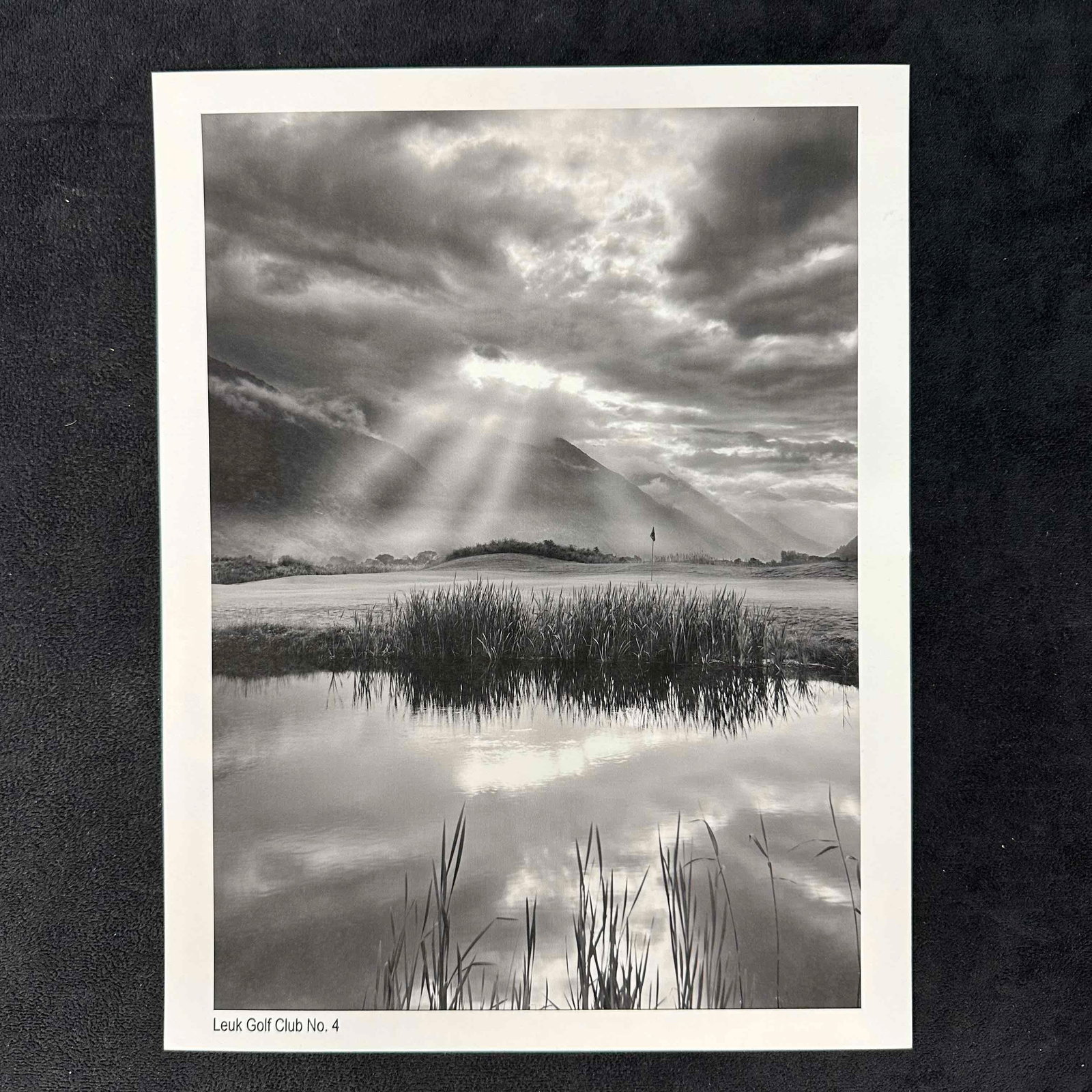 Leuk Golf Club No. 4 Black and White Golf Photography Print: Black-and-white photographic print of Leuk Golf Club’s No. 4 hole, featuring dramatic sun rays piercing through the clouds over a serene mountain backdrop. The foreground shows a reflective