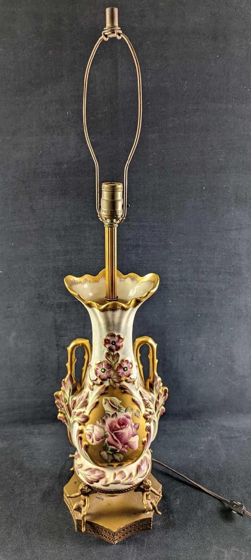Vintage Gilded Floral Porcelain Table Lamp: Vintage Gilded Floral Porcelain Table Lamp. This lamp looks like it was converted from a regular vase. Sits upon a brass-toned stand with cupids. The lamp is approx 8" x 8" x 34" (without the harp,
