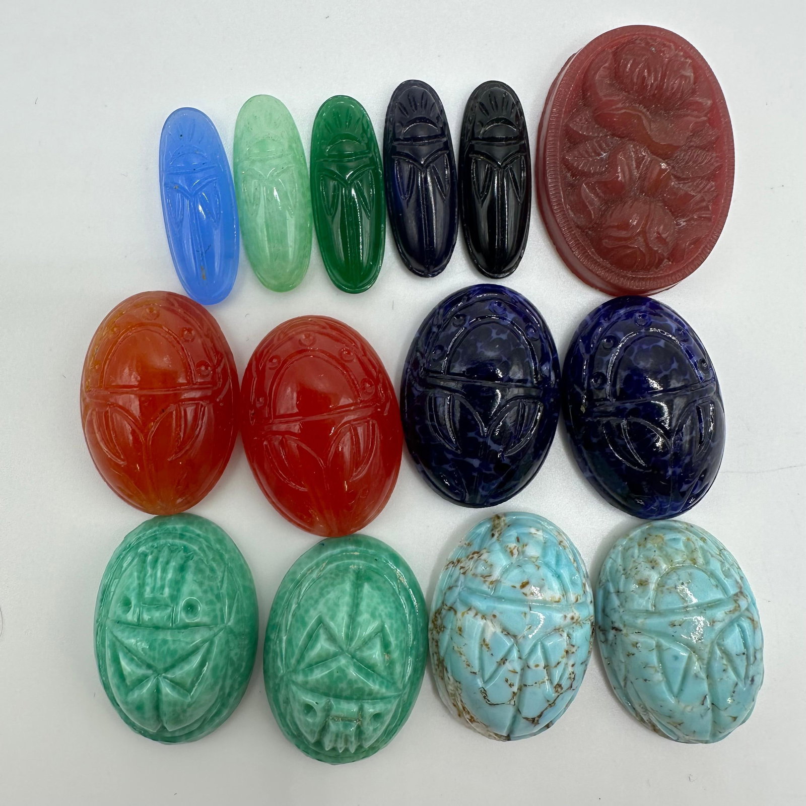 Carved Scarab Style Stone and Glass Cabochon Lot of 14: Group of assorted carved scarab-style cabochons in various colors, including green, blue, red, and black, along with one larger oval floral cameo-style piece. Materials appear to be mixed stone,
