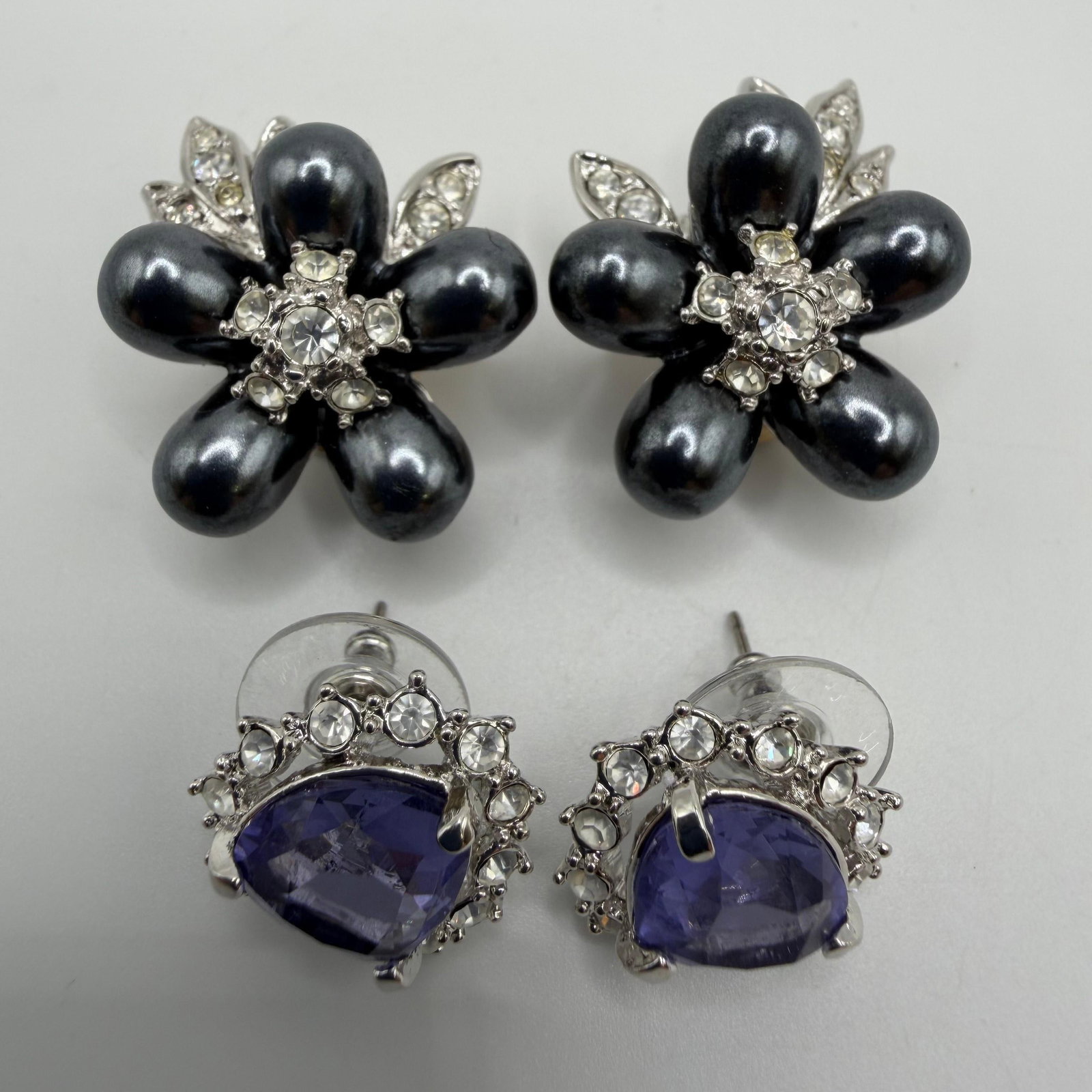 Nolan Miller Designer Costume Earrings Black Floral and Lavender Purple Set: Collection of two pairs of signed Nolan Miller designer costume earrings. Includes a pair of black floral clip-on earrings accented with clear rhinestones and silver-tone leaf details, along with a pa