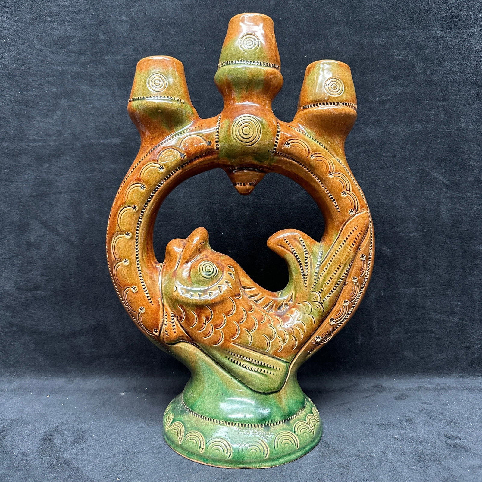 Art Pottery Figural Fish Three Neck Candlestick Holder: Art pottery vase featuring a figural fish within a circular form and three neck openings at the top, candlestick holders. Decorated with incised patterns and finished in a mottled green and brown glaz