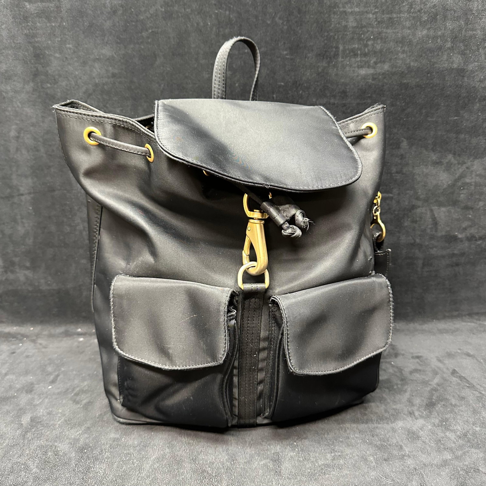 LJS Black Nylon Drawstring Backpack with Gold Tone Hardware: Black nylon backpack by LJS brand featuring a drawstring closure with flap top, gold-tone hardware, adjustable shoulder straps, dual exterior front pockets, and a zippered interior pocket. Interior ta