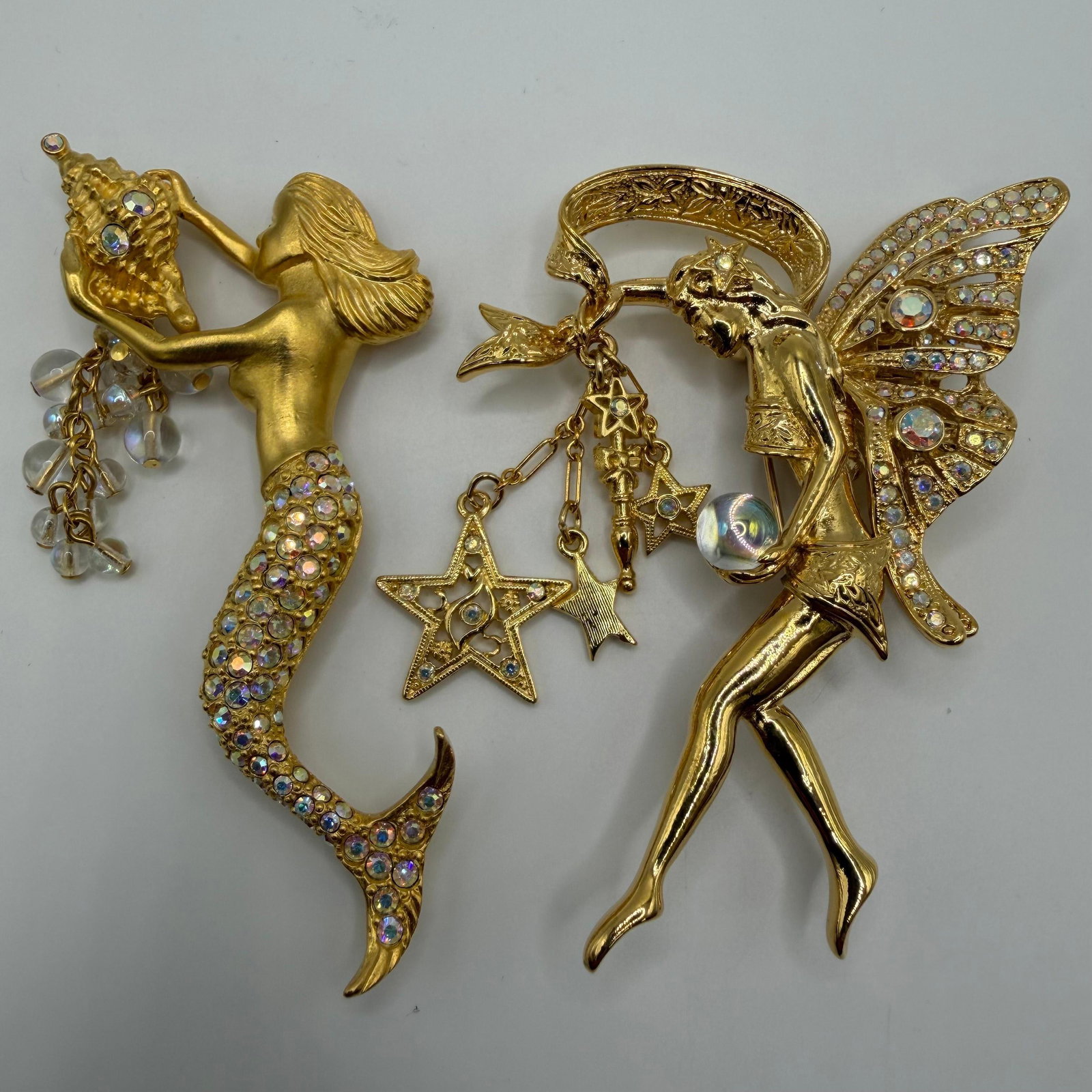 Pair of Kirks Folly Mermaid and Fairy Fantasy Brooches HH57: Matching pair of Kirks Folly gold-tone fantasy brooches, including a rhinestone mermaid holding a jeweled seashell with dangling iridescent bead accents, and a fairy with rhinestone-set wings holding