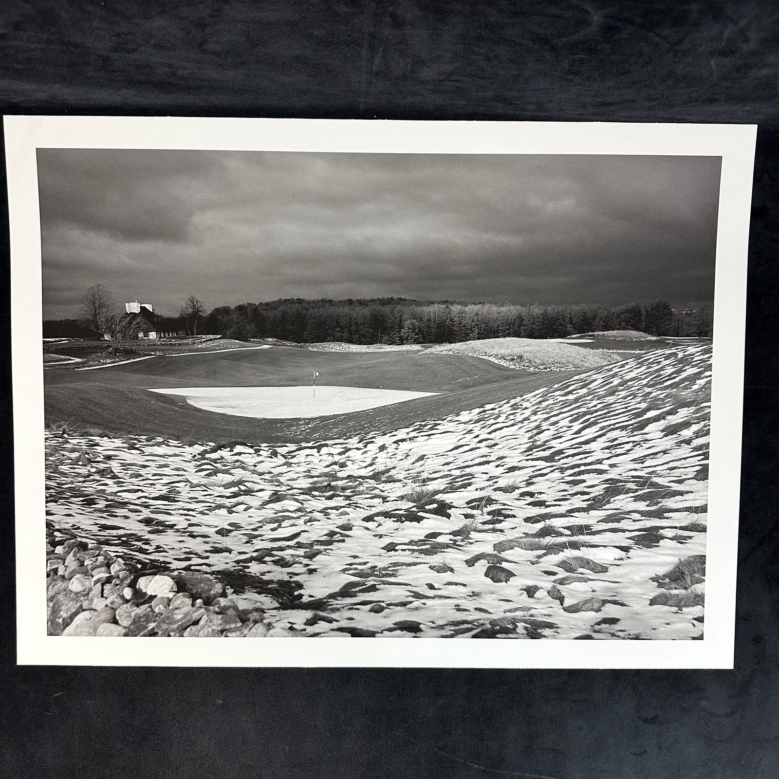Snow-Dusted Golf Green with Clubhouse in Distance Inkjet Photo Print: Black-and-white inkjet photographic print depicting a striking golf course landscape in winter. The scene shows a green surrounded by rolling hills with patches of snow scattered across the ground. A 