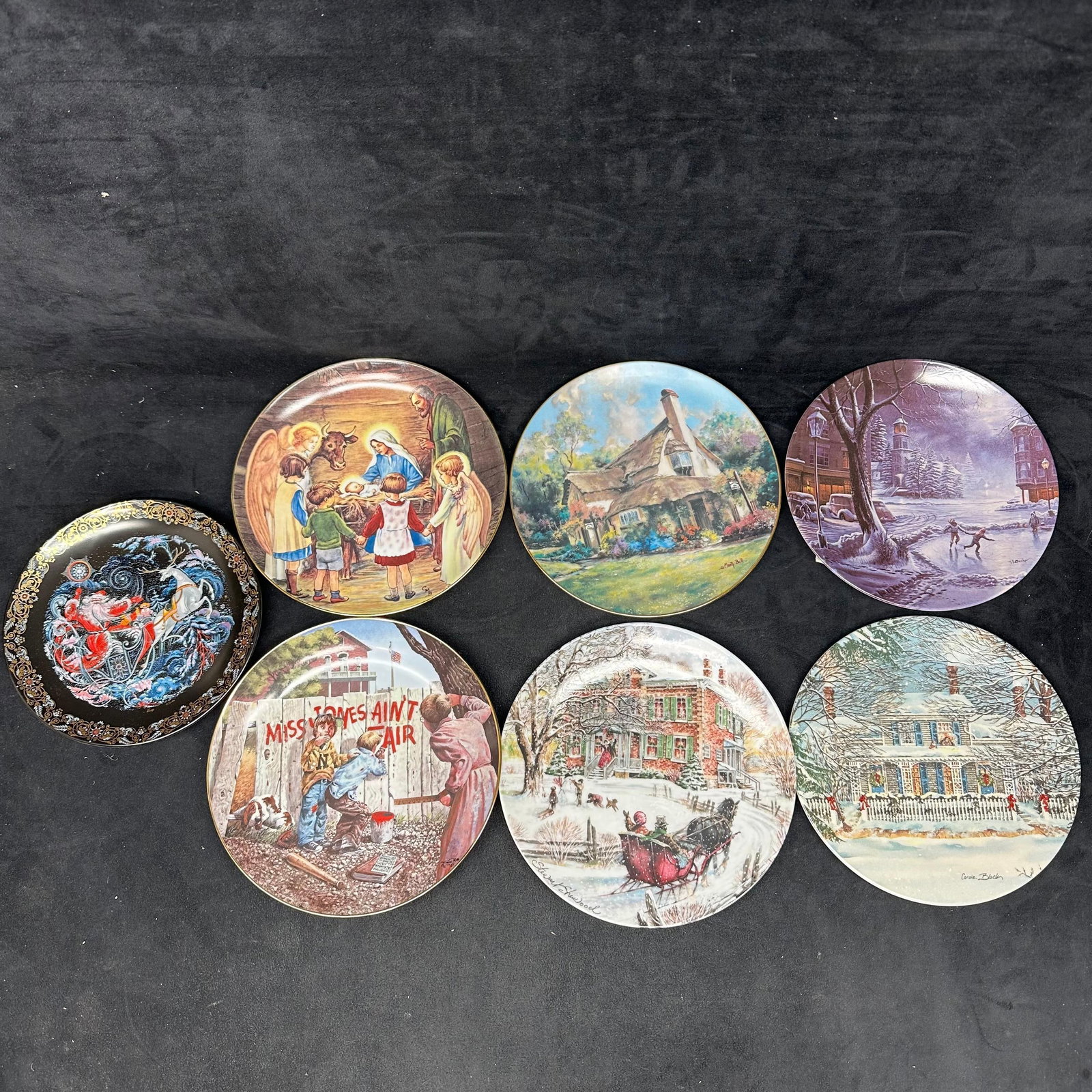 Collection of Seven Collector Plates Everyday and Winter Scenes: Group of seven decorative collector plates featuring everyday cottage, village, and winter scenes by various artists. Plates display seasonal imagery, including snow-covered villages, illuminated home