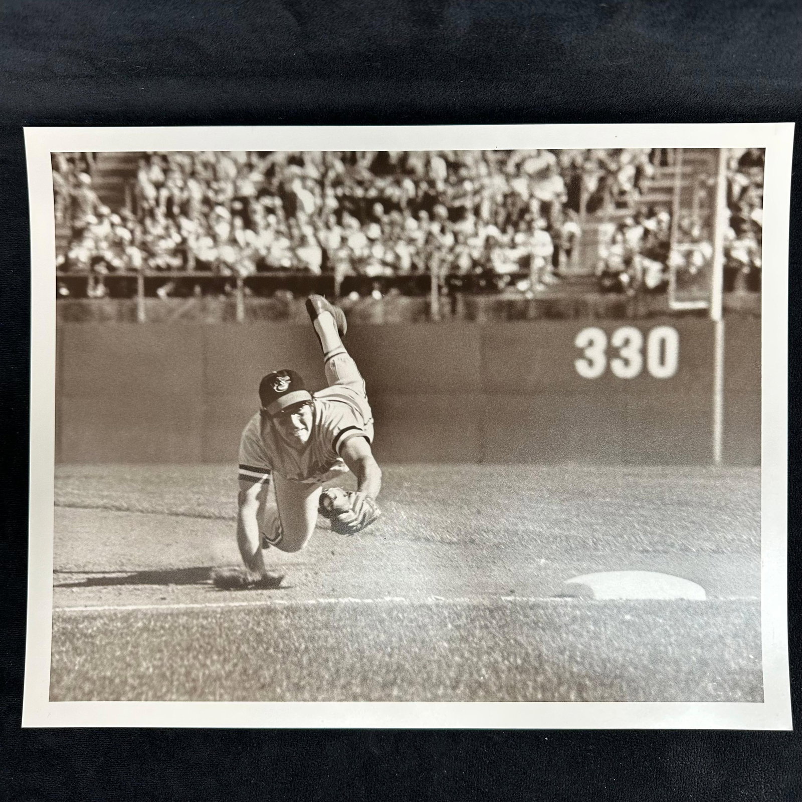 Analog Photograph Print Baltimore Orioles Player Diving Catch Black and White: Black and white analog photo print capturing a Baltimore Orioles player mid-air while making a diving catch near third base. Stadium crowd and "330" outfield marker visible in the background. 