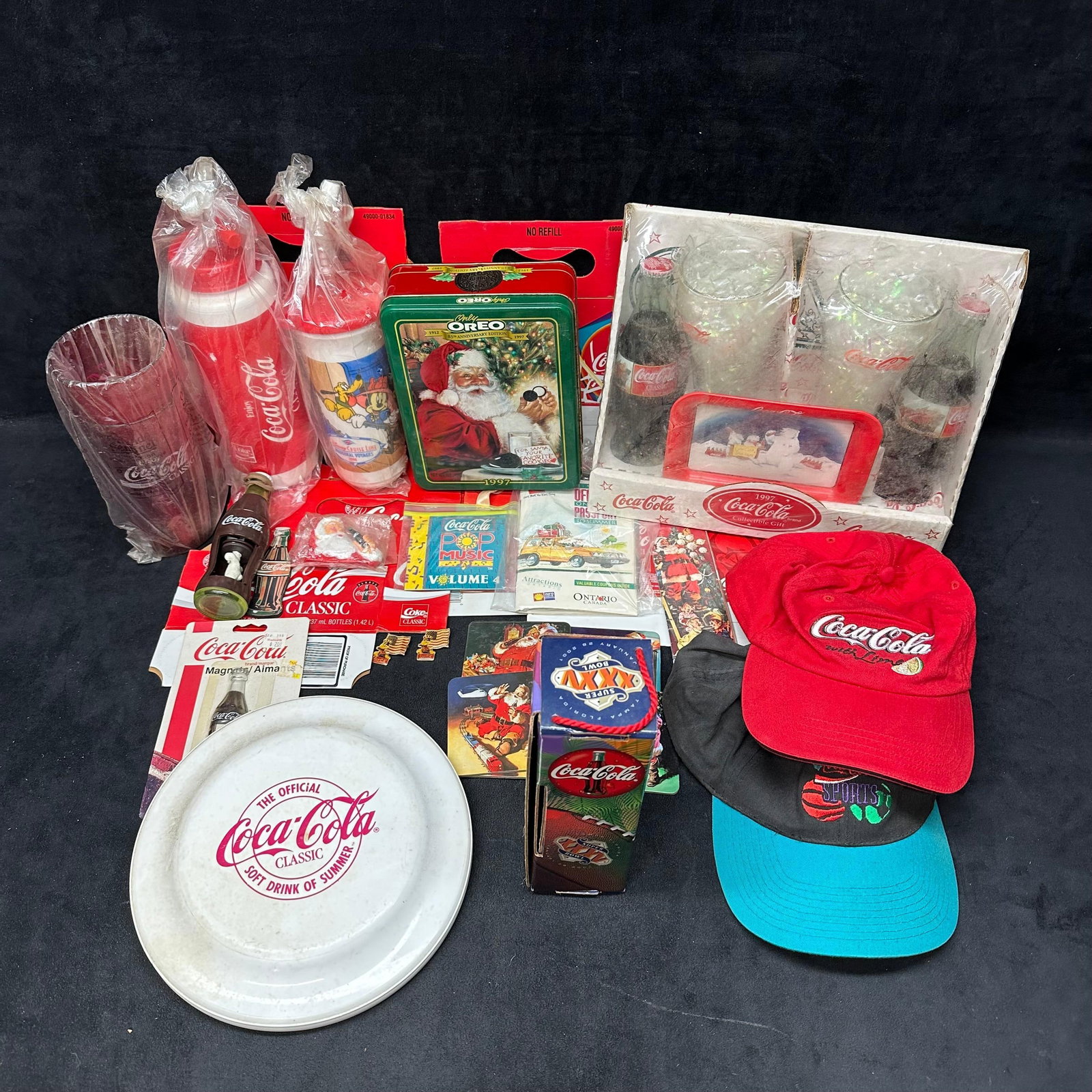 Coca Cola Collectibles Lot Bottle Glassware Hats Tin Paper Lot: Large mixed lot of Coca-Cola collectibles, primarily 1990s era. Includes boxed and loose items such as glassware sets, refillable drink bottles, small glass bottles, serving tray, tin, magnets, pins, 