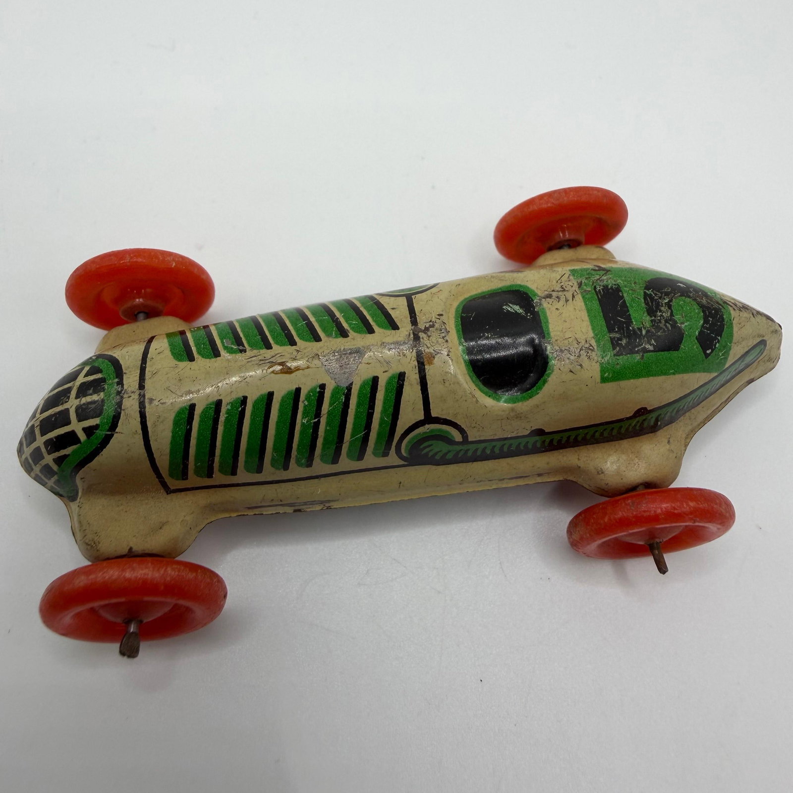 Vintage German Tin Lithograph Race Car Toy: Vintage Made in Germany tin lithograph racing car featuring cream, green, and black graphics with four red wheels. Pressed tin construction with simple axle design. Shows surface wear, scratches, pain