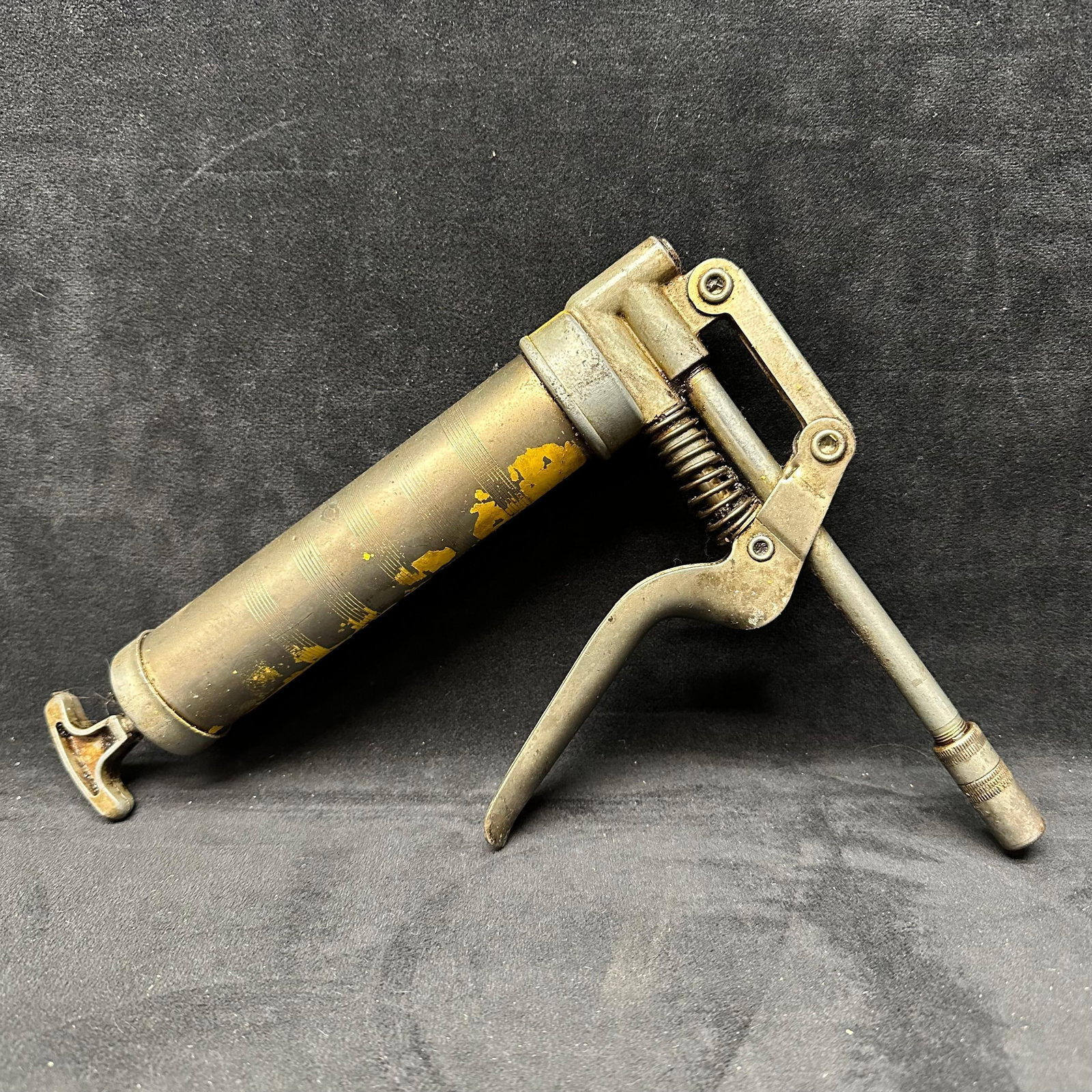 Vintage Pistol Grip Grease Gun Metal Body: Vintage pistol-grip style grease gun constructed of metal with spring-loaded trigger mechanism and rigid nozzle. Features a cylindrical barrel with remnants of yellow paint and knurled fittings. Exhib