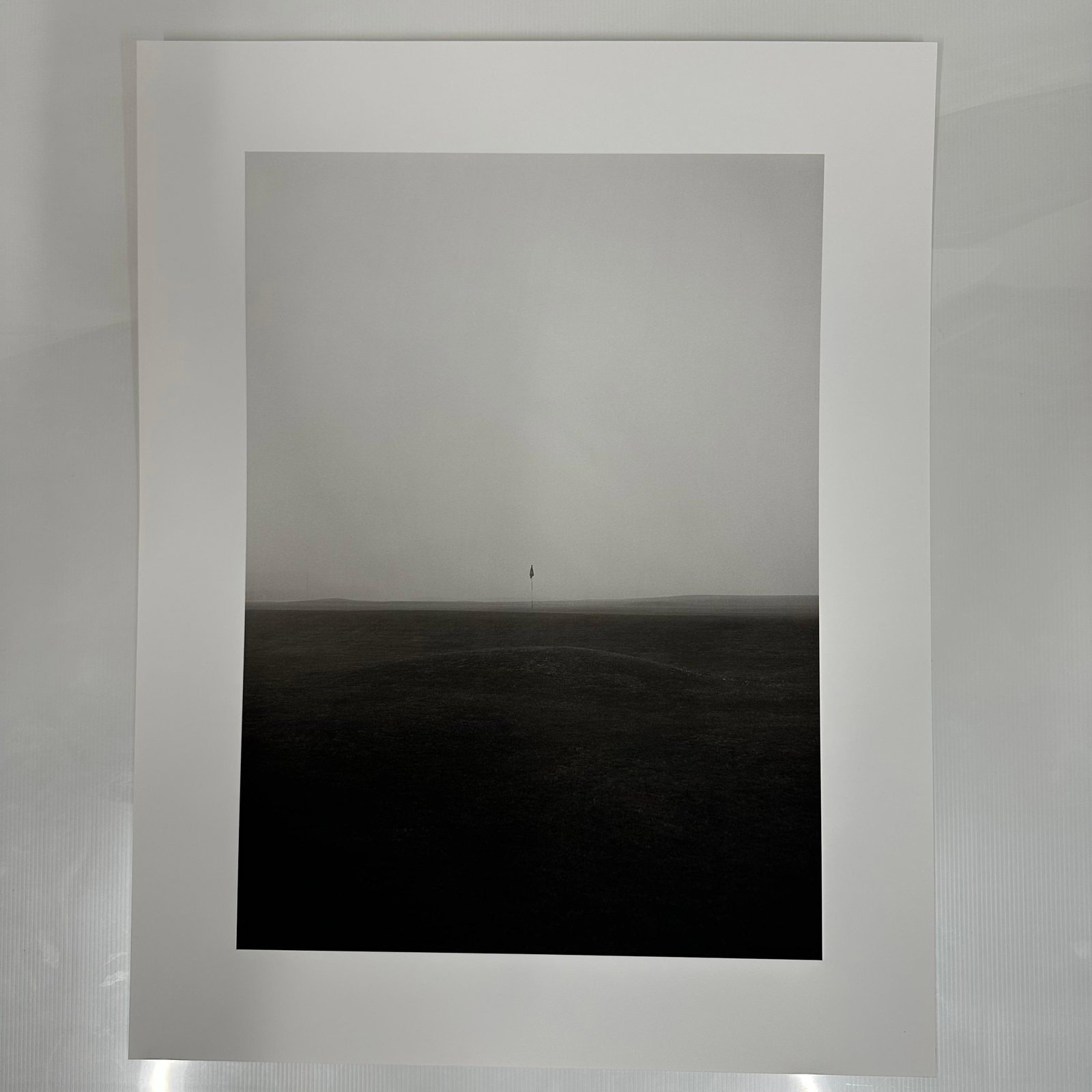 Black and White Minimalist Golf Course Photography Print Foggy Fairway Scene #3: Black and white photographic print depicting a solitary golf flag standing on a mist-covered fairway. The minimalist composition and muted tones create a serene, almost abstract landscape. Printed on 