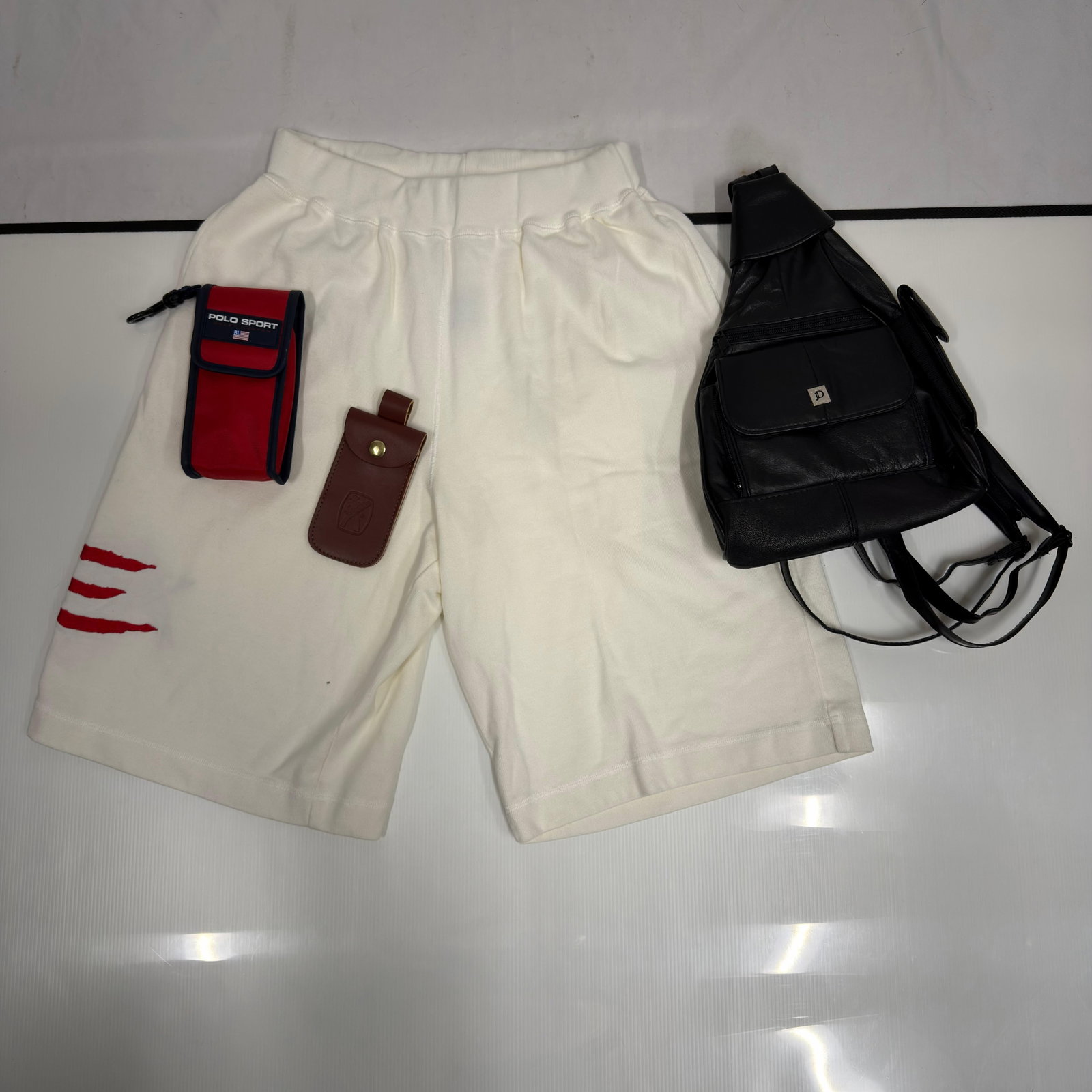 Nautica Sailing USA Shorts and Mens Bags Accessories Lot: Mixed men’s fashion lot including vintage-style Nautica Sailing USA white sweat shorts with an embroidered American flag design, Polo Sport Ralph Lauren red utility pouch, brown leather accessor