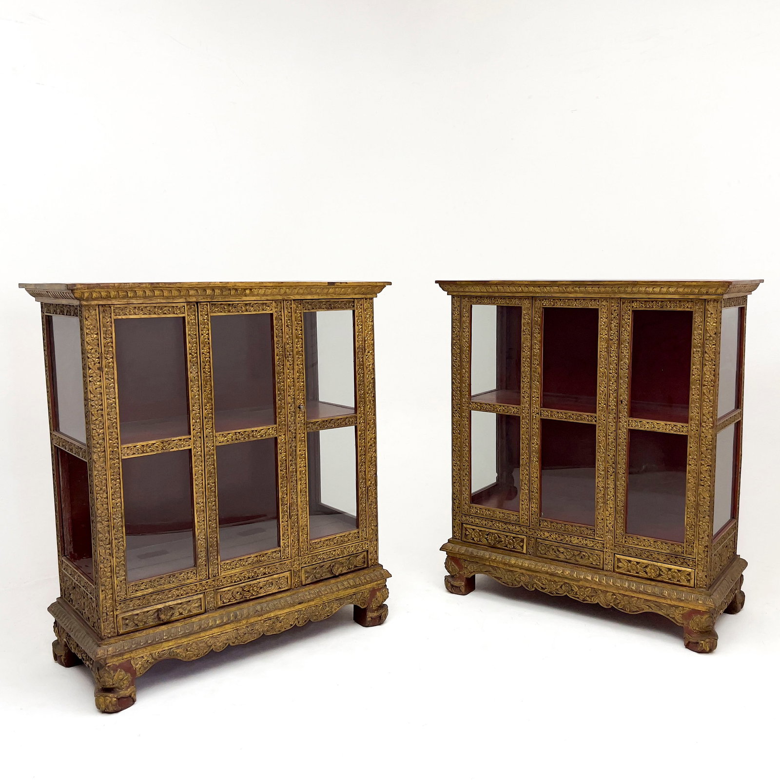 A Pair of Thai Giltwood Vitrines: Lot 46 A Pair of Thai Giltwood Vitrines Hardwood, glass, paint, gilt lacquer each: 49 1/2 x 43 1/2 x 20 1/2 inches Description: A pair of 20th-century Thai traditional style vitrines heavily carved