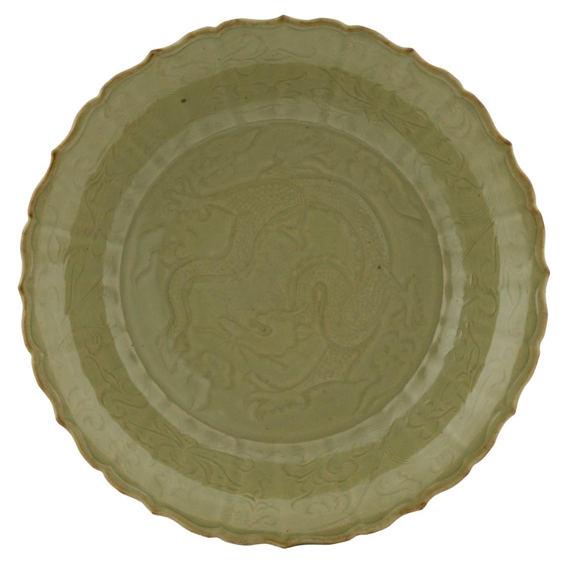 A LARGE CARVED DRAGON MING LONGQUAN CELADON CHARGER: Chinese Ming Dynasty LongQuan celadon barbed-rim charger. Shallow body, with a slightly flared lip with a shallow draft. Centrally festooned with a dynamic dragon soaring amidst flames and vapors. The