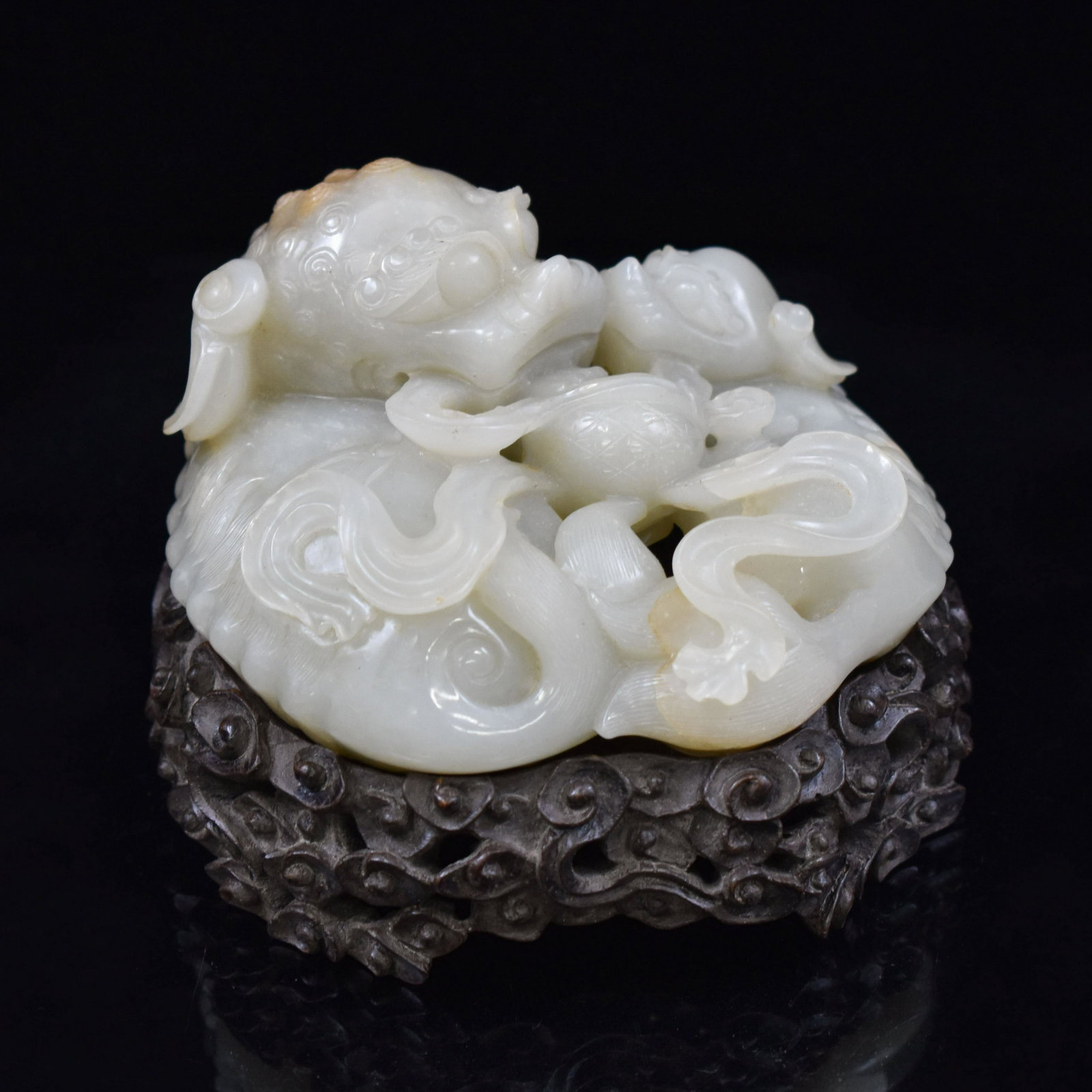 CARVED WHITE JADE OF THREE FOO-DOGS ON STAND: Measurements: Measurements (on stand): 98.425mm H x 92.075mm W Weight (Jade): 331.8g Fine Carved Chinese white jade recumbent three Foo-Dogs, its mouth holding a branching leaves, while one of smaller